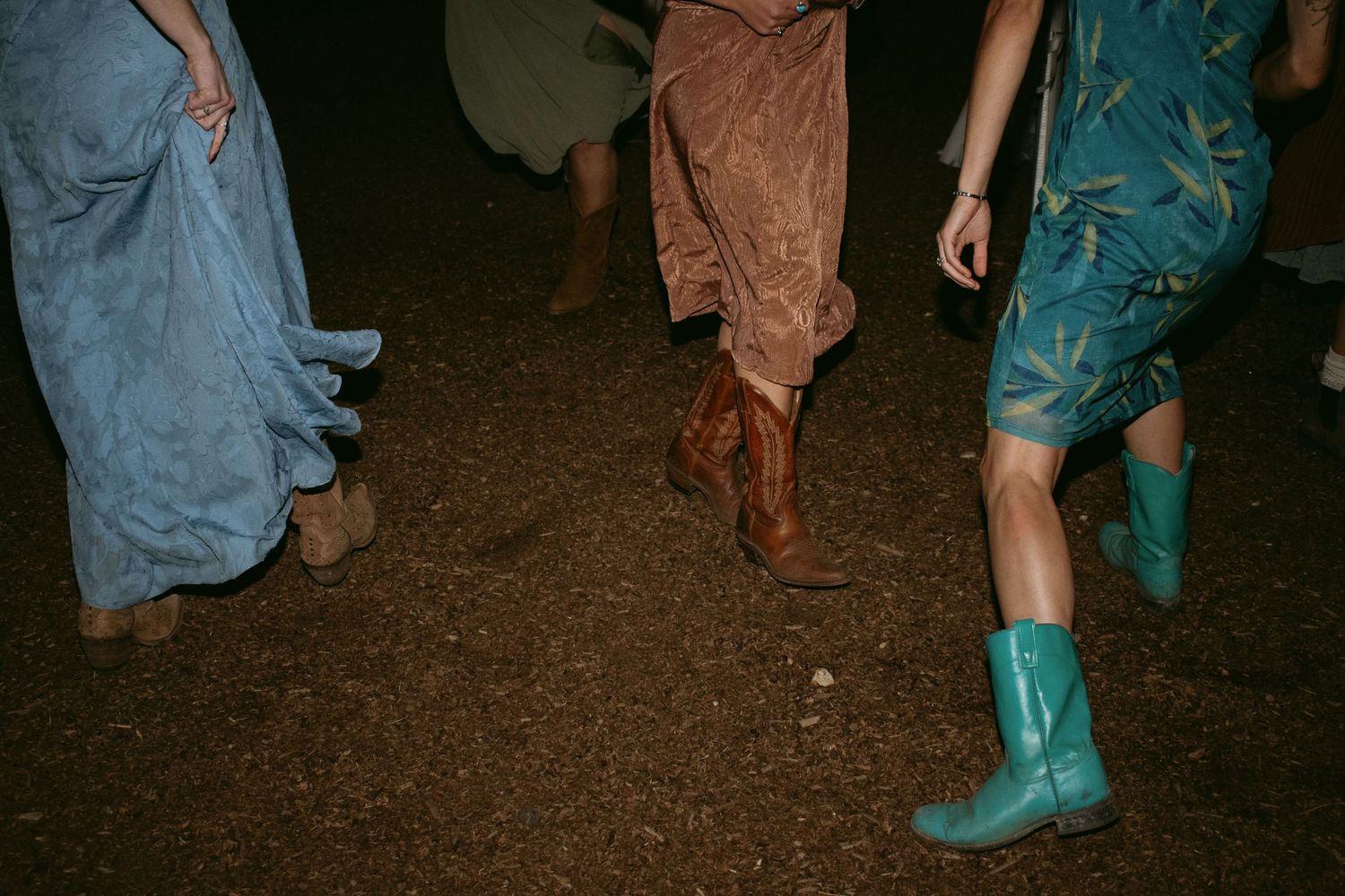 Three women wearing colorful dresses and cowboy boots dancing outdoors at night on dirt ground.