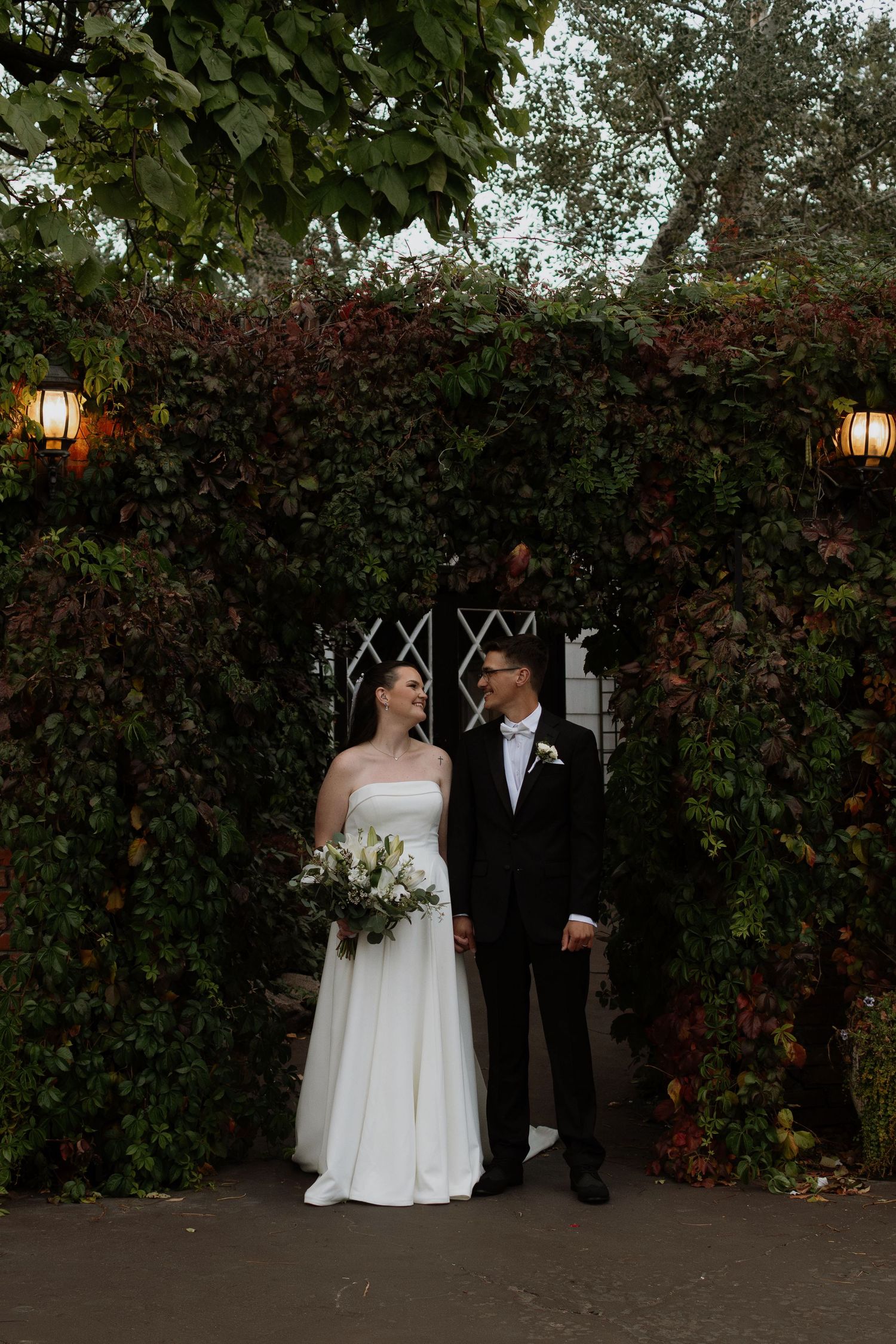 Bride and groom stand hand in hand beneath an ivy-covered archway with warm lanterns at their wedding venue.