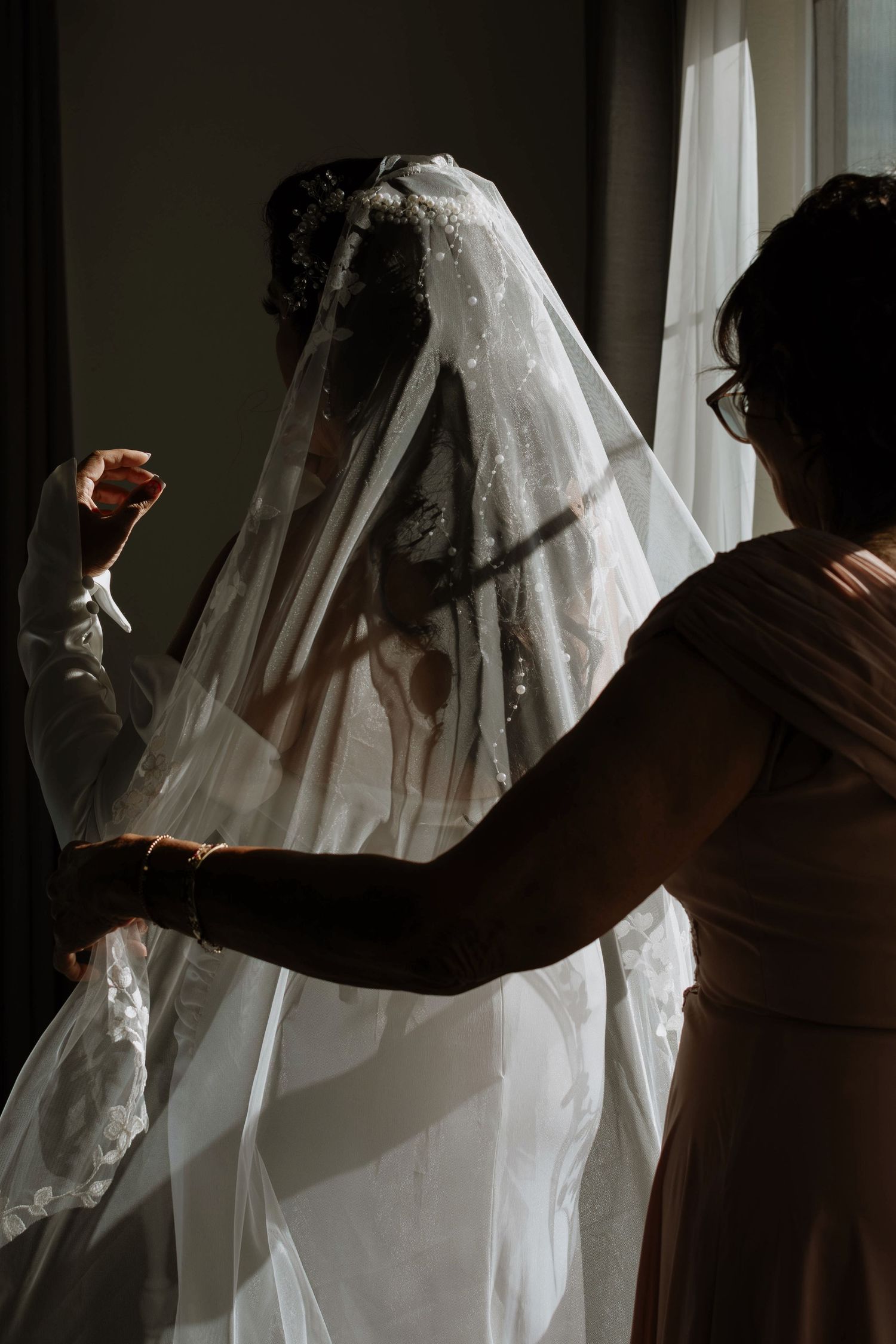 Silhouette of bride in wedding dress and veil being adjusted by bridesmaid near sunlit window.
