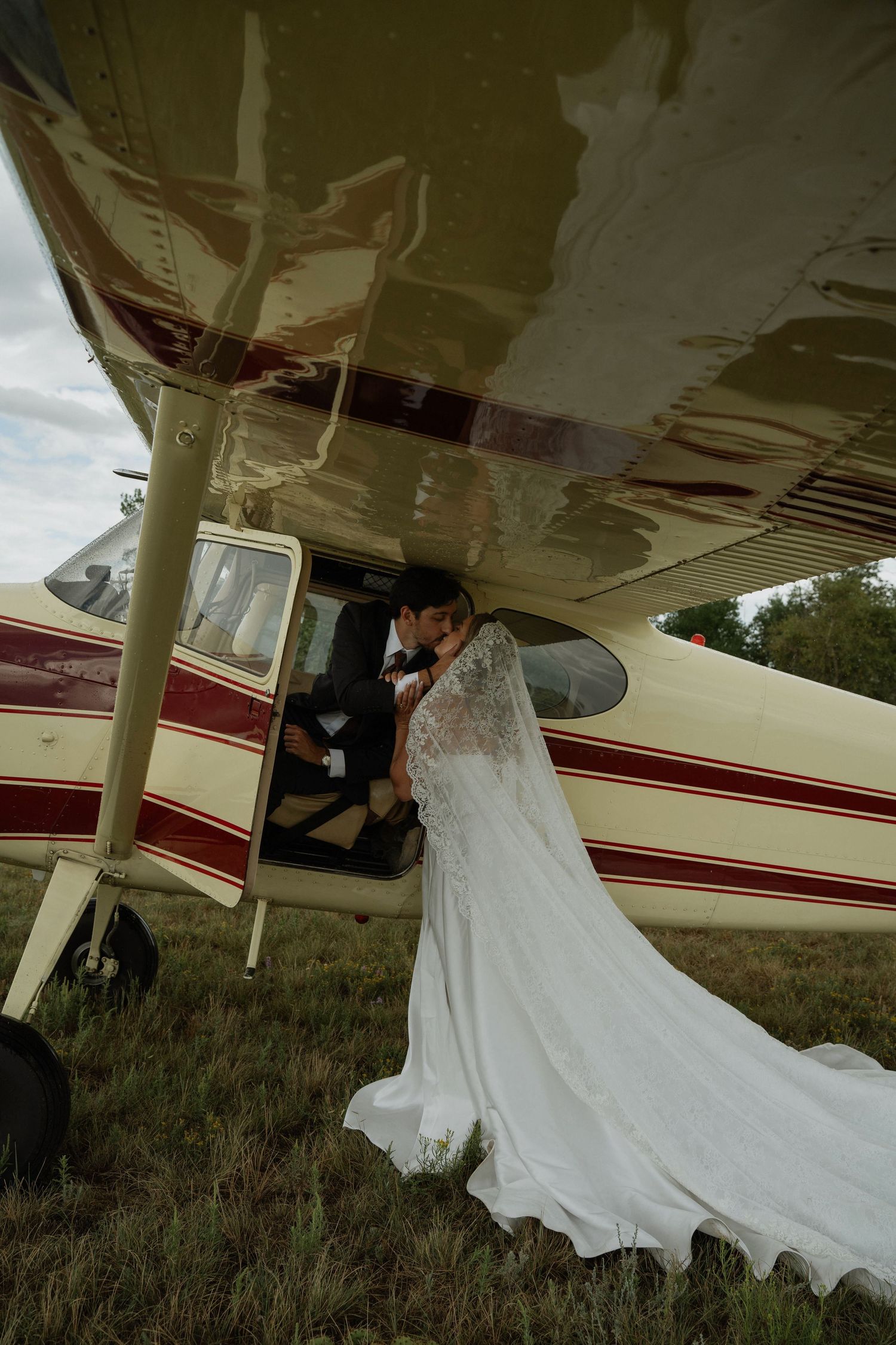 Bride in white gown kisses groom seated in small vintage plane door on grassy airfield.