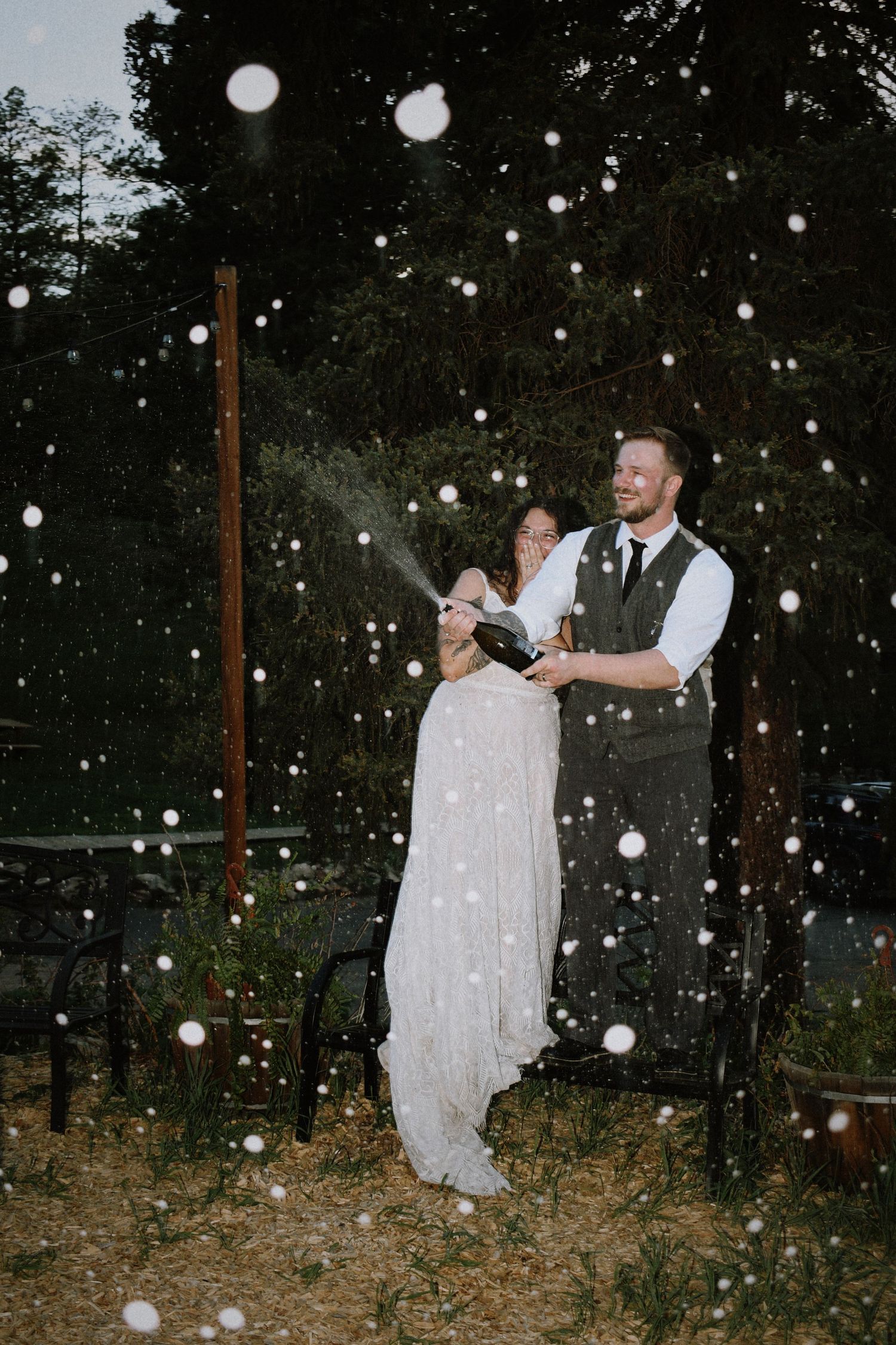 Joyful bride and groom popping champagne bottle with festive bokeh lights at outdoor evening wedding celebration.