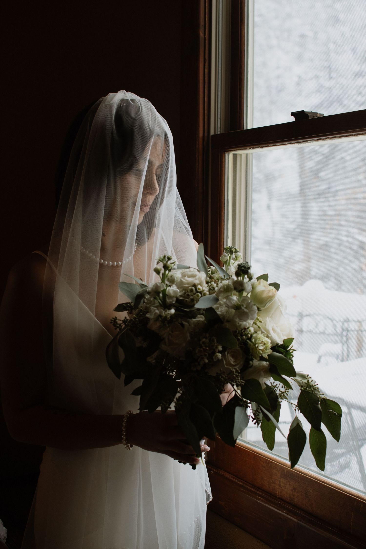 Bride wearing veil and pearl necklace holding white floral bouquet while standing by a snow-covered window.