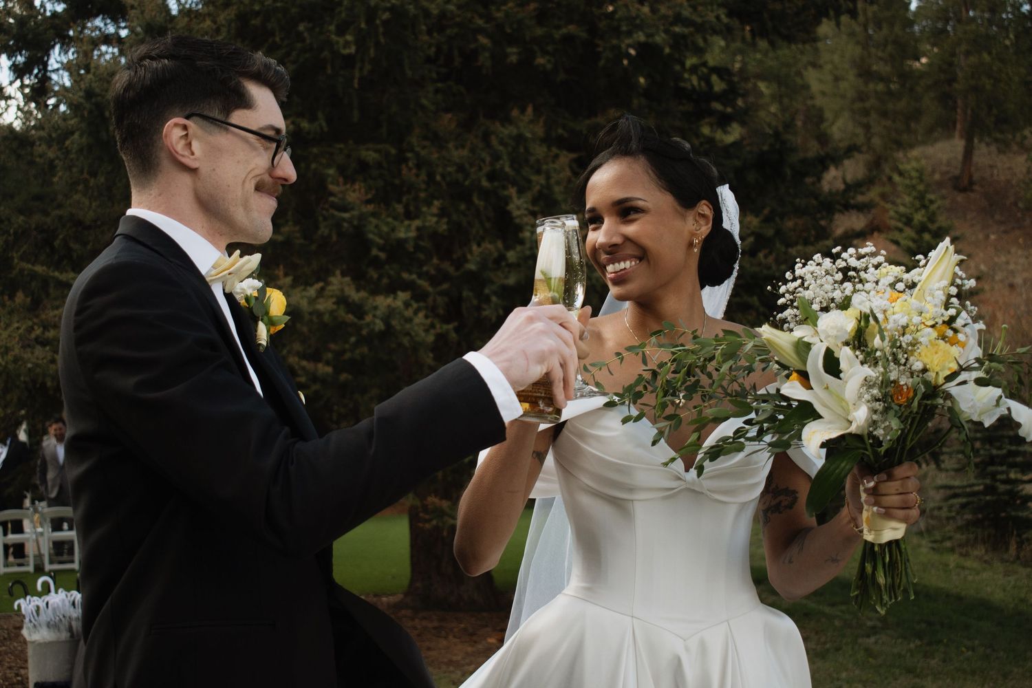 Bride and groom toasting with champagne glasses outdoors, bride holding white floral bouquet in elegant wedding attire.