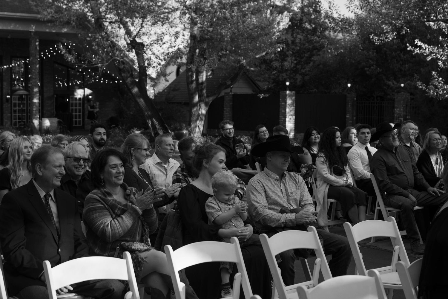 Crowd of guests seated in white chairs outdoors at an evening event with string lights in black and white photo.