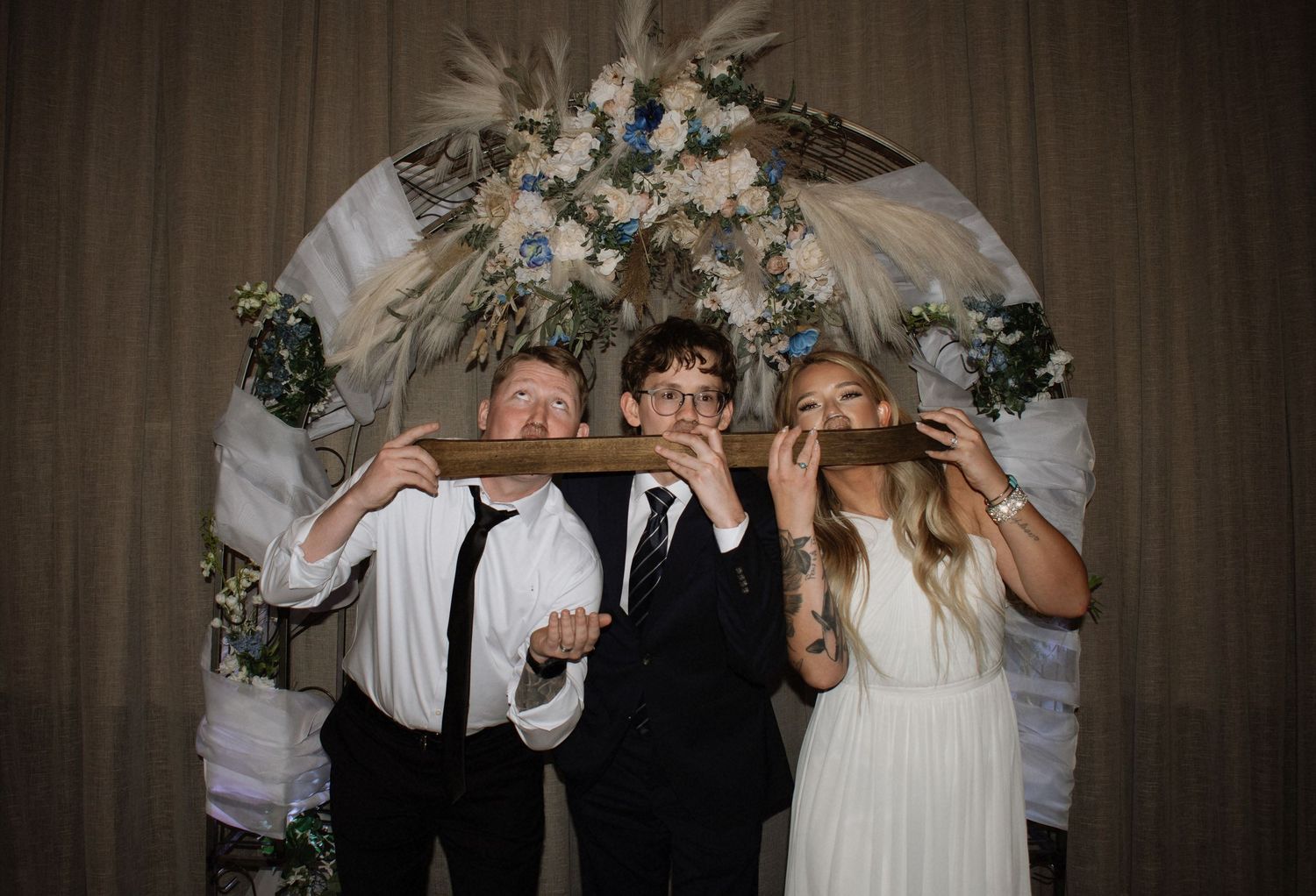 Three wedding guests pose playfully holding a wooden sign under a floral arch with pampas grass and blue flowers.