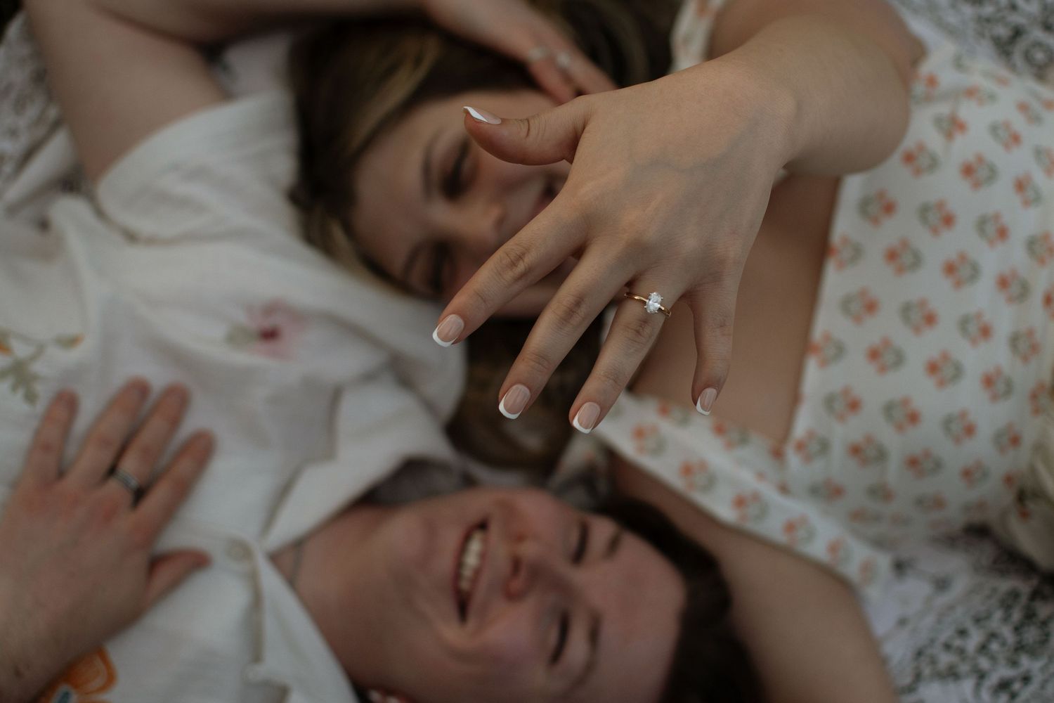 Two women lying down laughing, one showing off a diamond engagement ring on her hand.