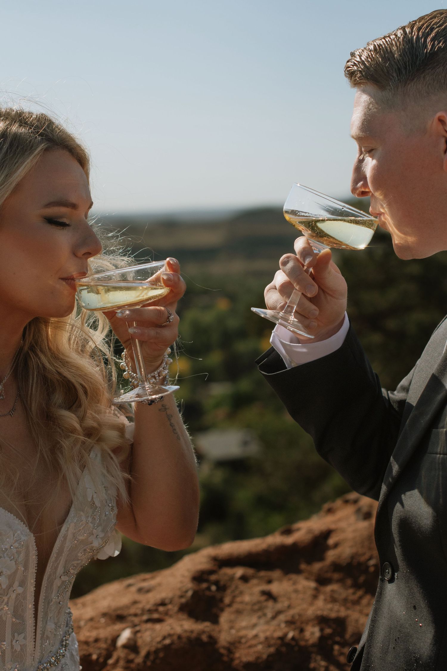 Bride and groom toast with champagne glasses at outdoor wedding celebration with scenic backdrop.