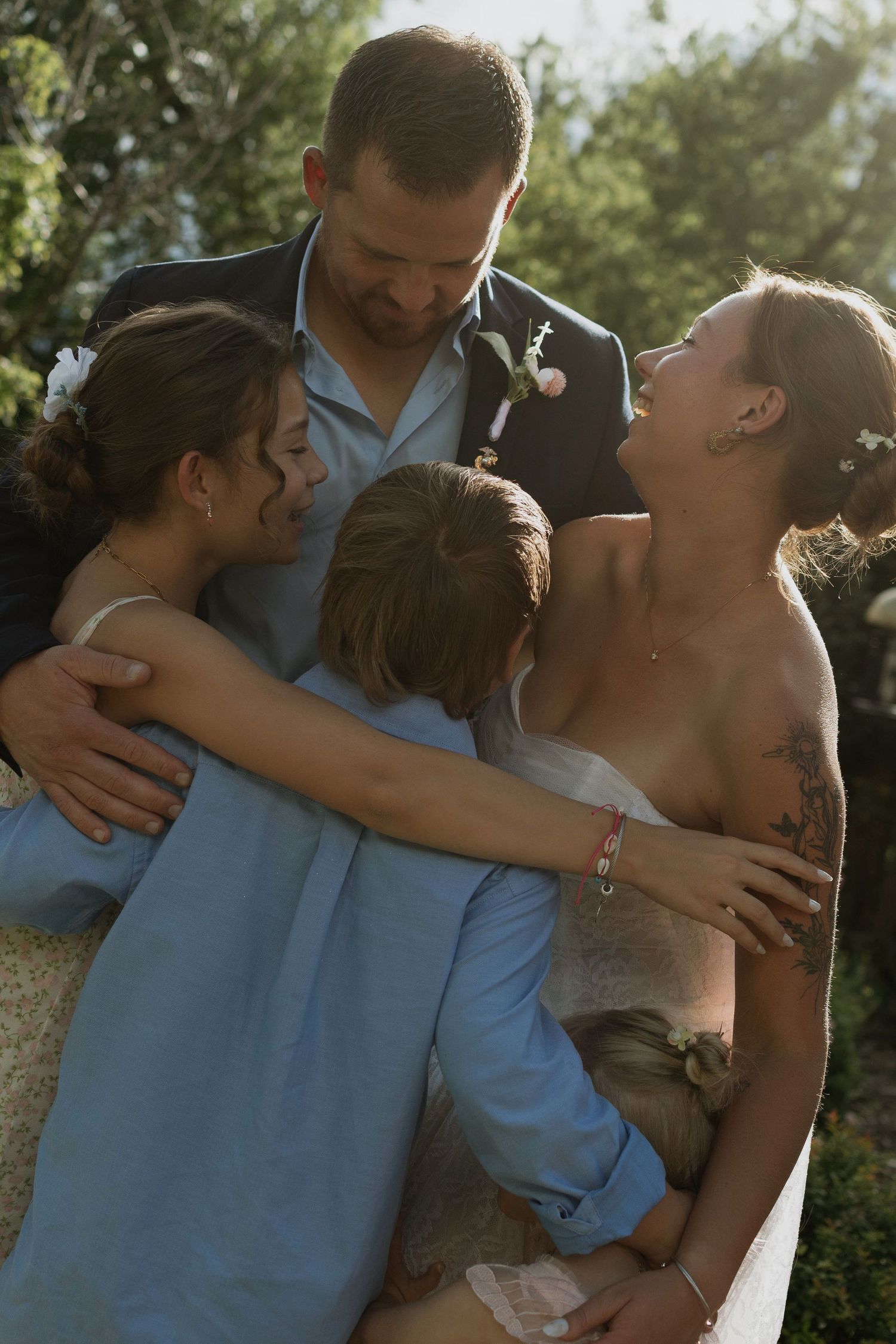 Newlyweds share a warm group hug with children in golden outdoor light at a wedding celebration.