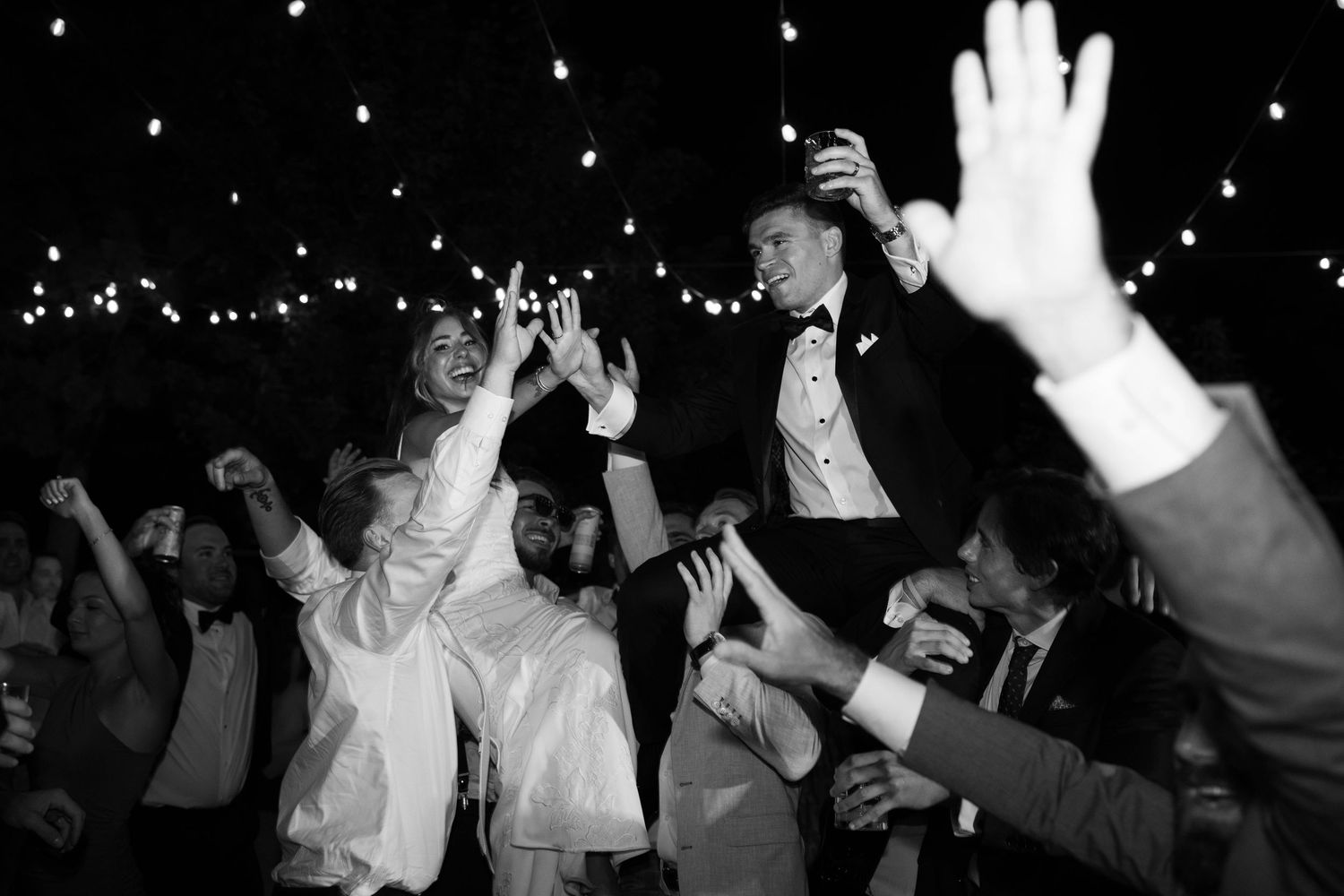 Black and white wedding reception photo of bride and groom being lifted by guests while dancing under string lights.