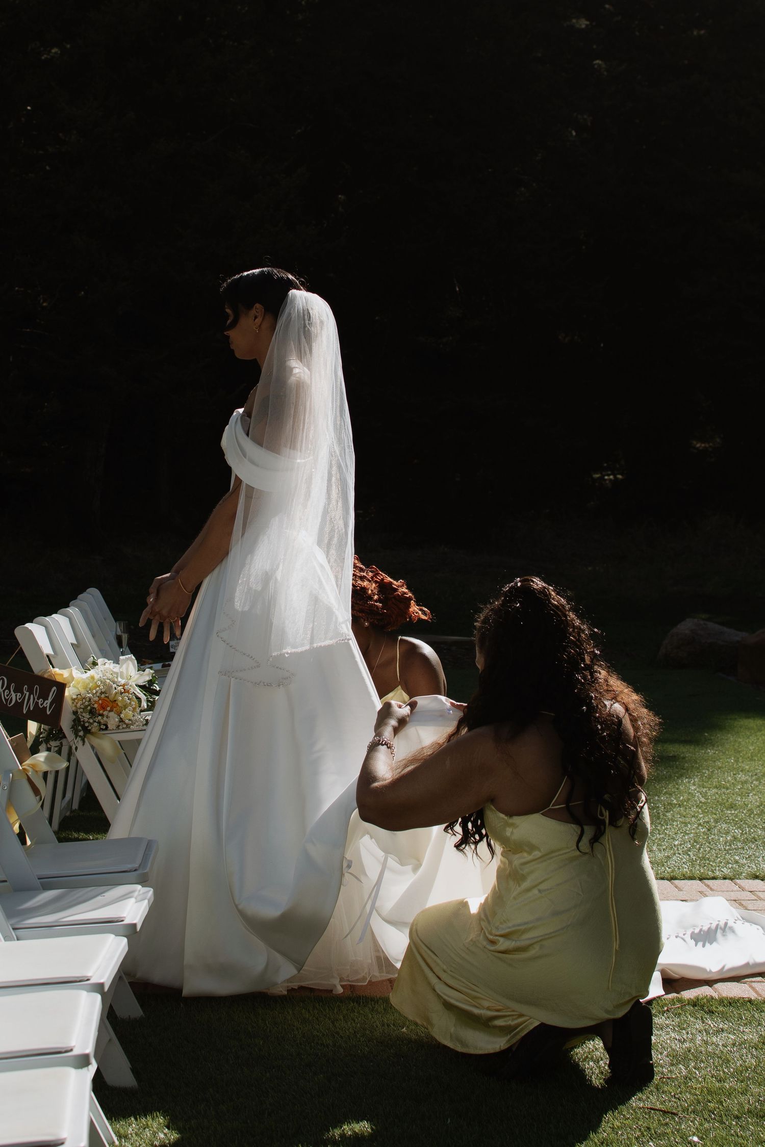 Bride in white gown and veil being assisted by bridesmaid in yellow dress at outdoor wedding ceremony.