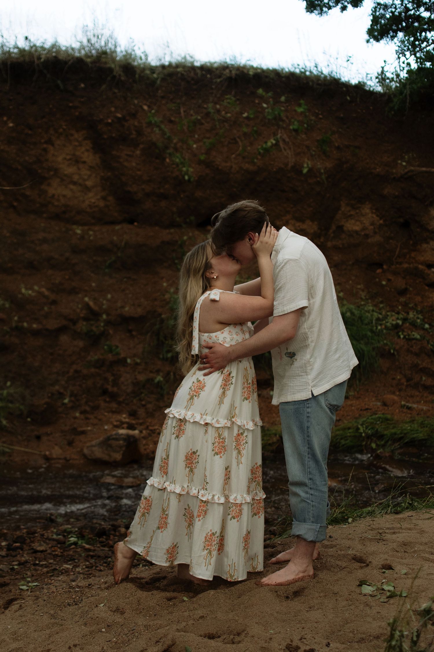 Couple sharing a romantic kiss outdoors on sandy ground with an earthy hillside backdrop, woman in floral dress.