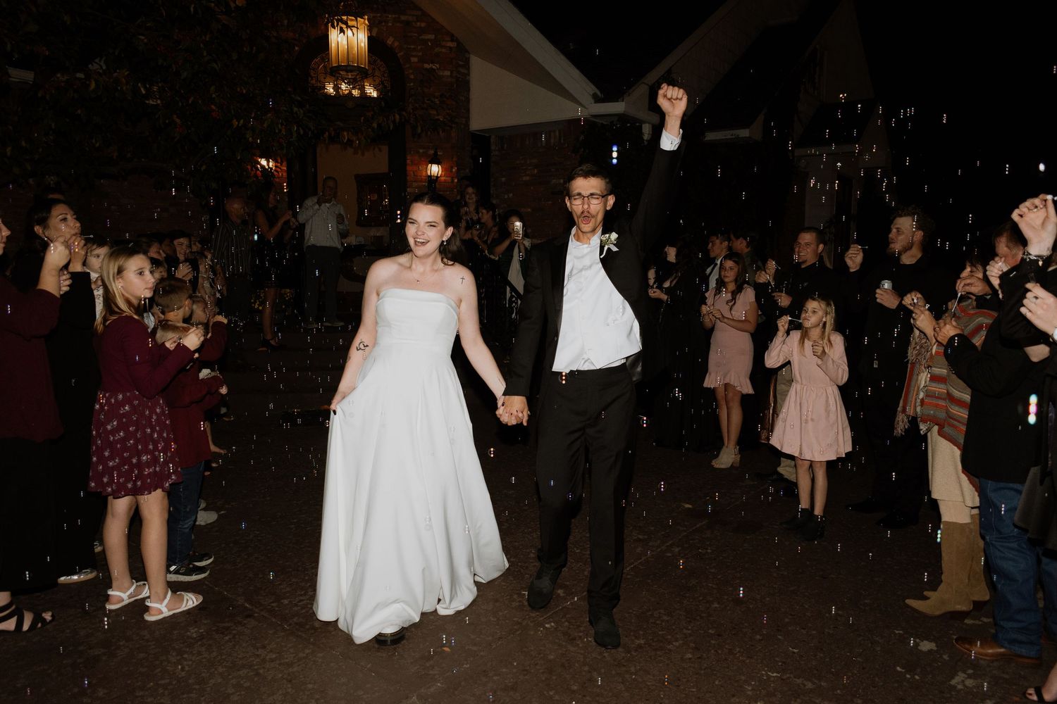 Bride and groom exit wedding reception holding hands, groom raising fist in celebration, surrounded by cheering guests.
