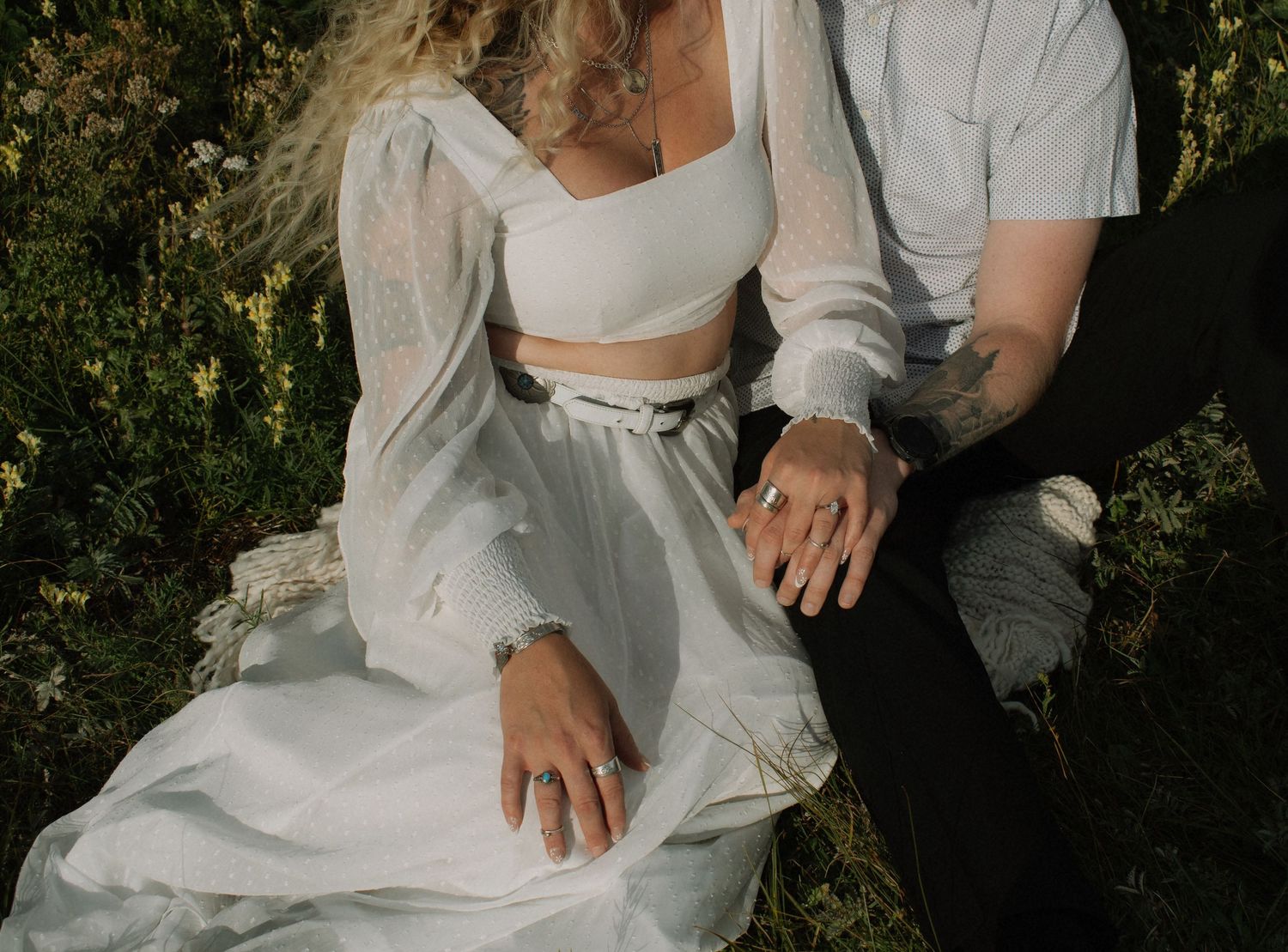 Couple in white outfits sitting in a flower field, showing off engagement rings and tattoos.