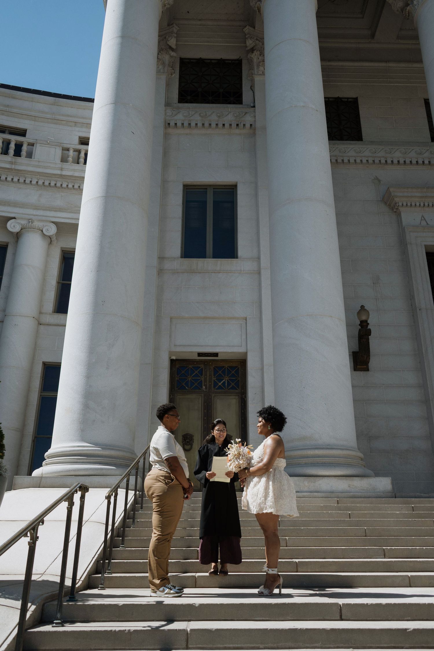 Couple exchanges vows with officiant on steps of a grand white columned building on a sunny day.