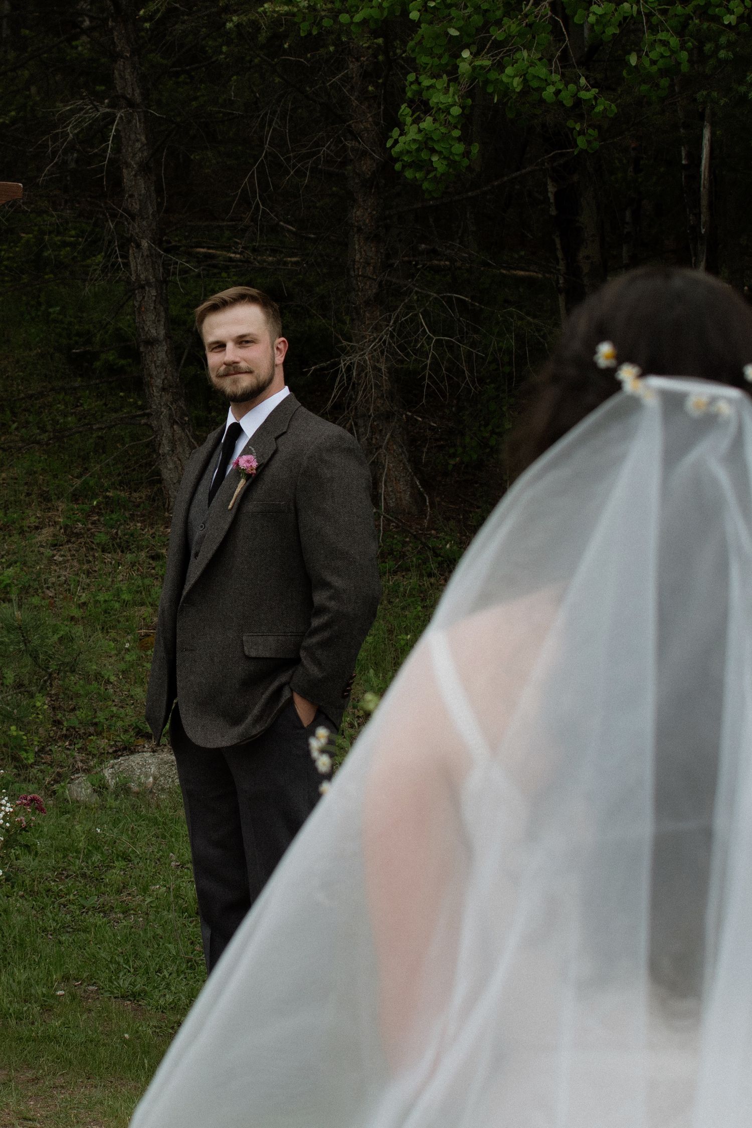 Groom in gray suit smiles at bride in white veil during outdoor first look wedding moment in forest.