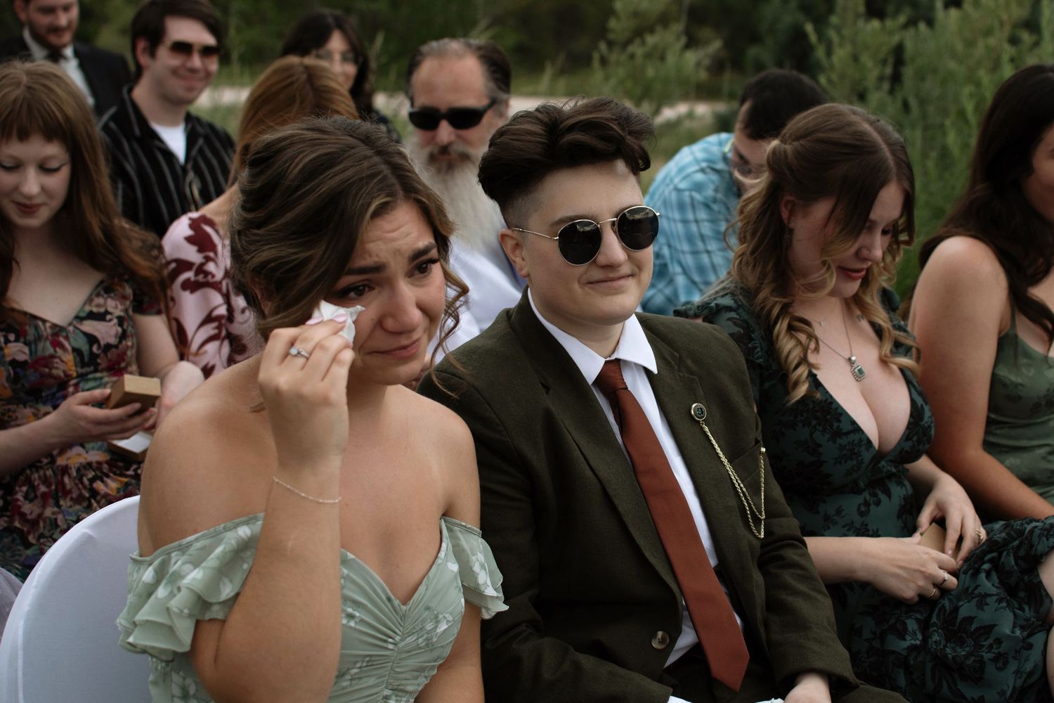 Emotional wedding guests seated outdoors, woman wiping tears, man in sunglasses and suit, during outdoor ceremony.