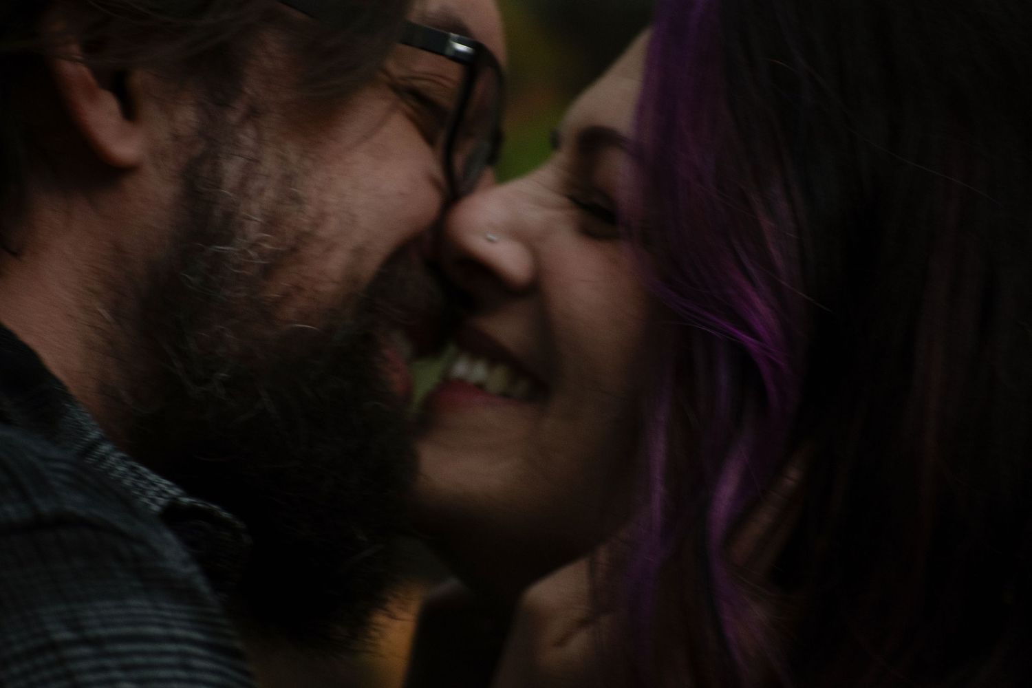 A couple with purple hair and glasses share a romantic close moment, smiling tenderly at each other.