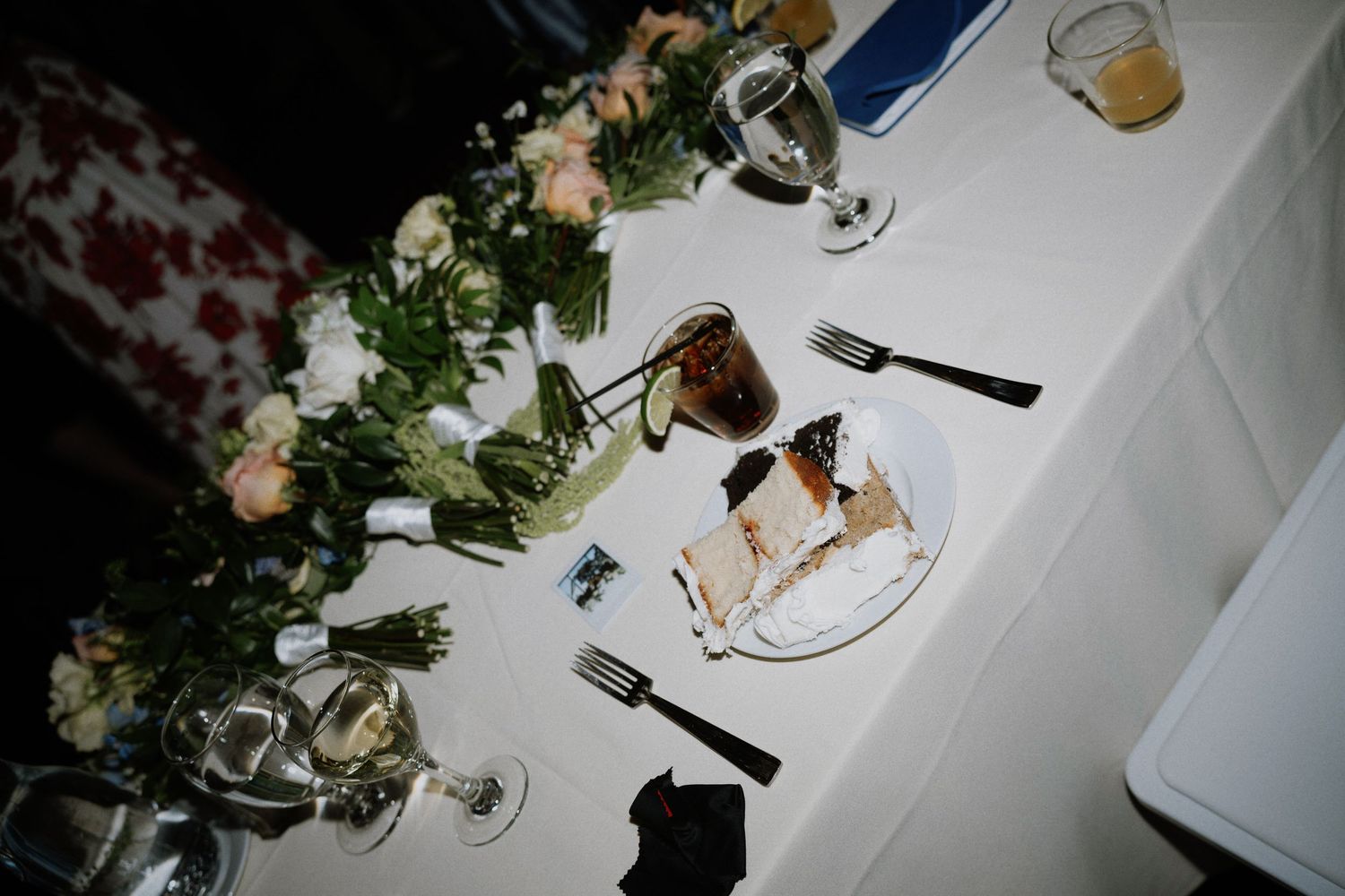 Elegant wedding reception table with floral centerpiece, wine glasses, and a slice of wedding cake on a white tablecloth.