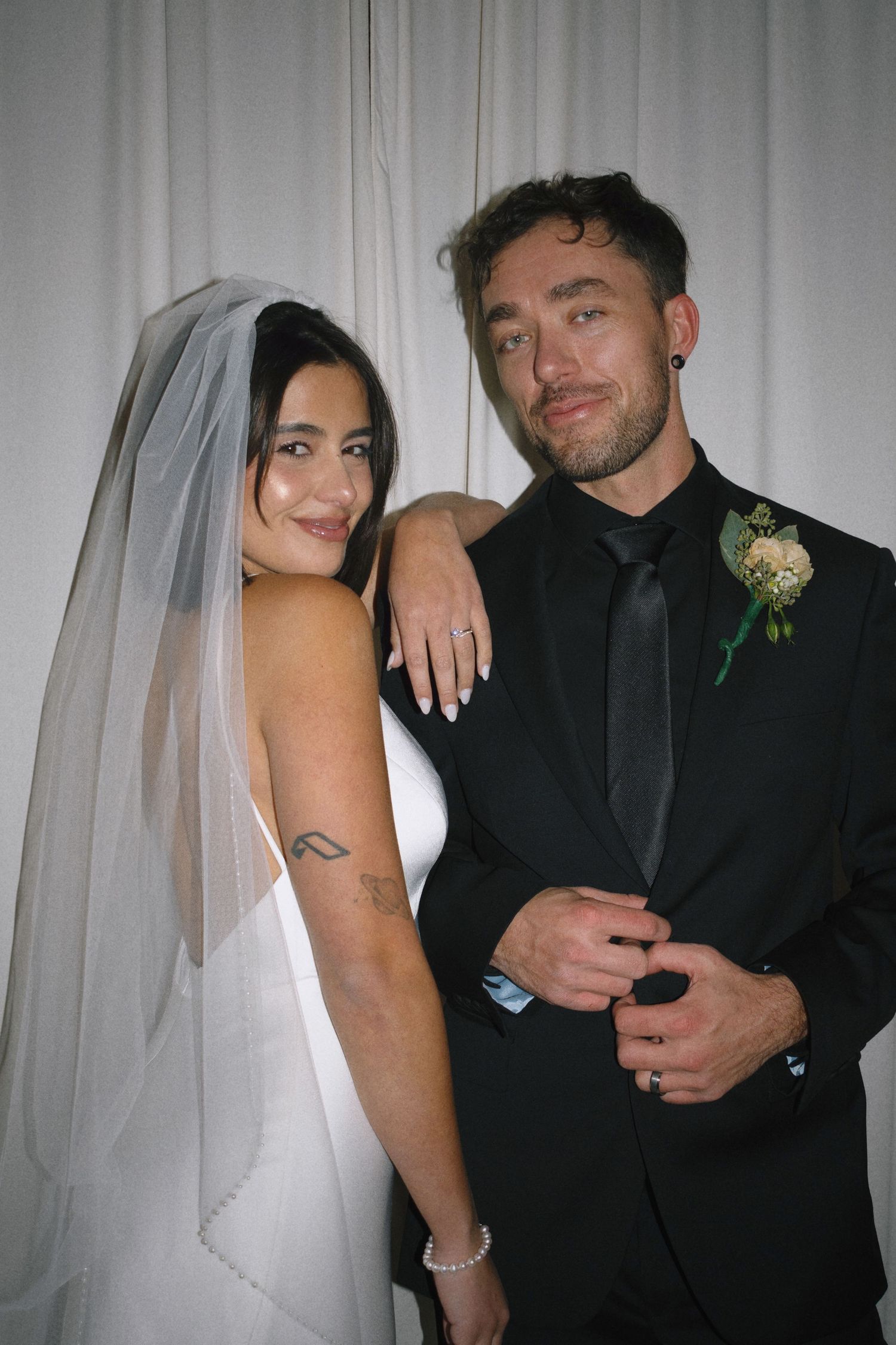 A bride in white dress and veil poses with groom in black suit and boutonniere against white curtain backdrop.