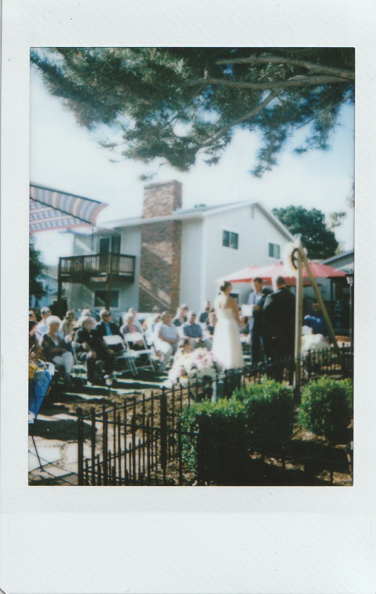 Outdoor wedding ceremony in a backyard garden with guests seated in white chairs near a house on a sunny day.
