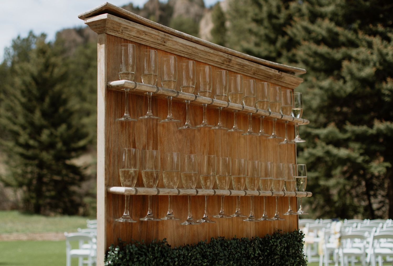 Rustic wooden champagne glass wall display rack outdoors at a wedding venue with white chairs and trees in background.