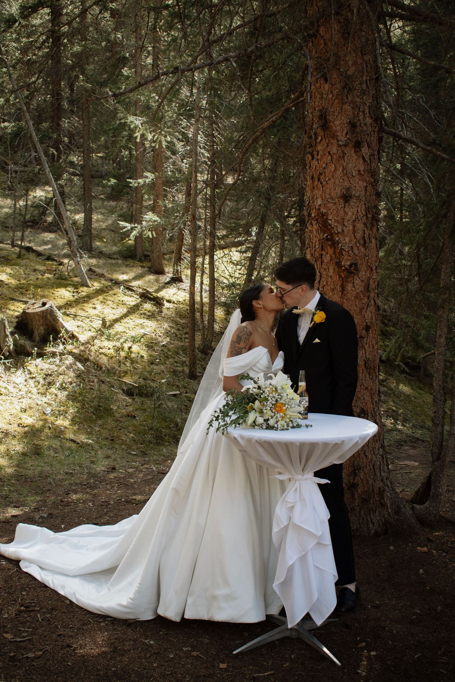 Bride and groom share a kiss beside a tall pine tree in a sunlit forest, with a cocktail table and bridal bouquet nearby.