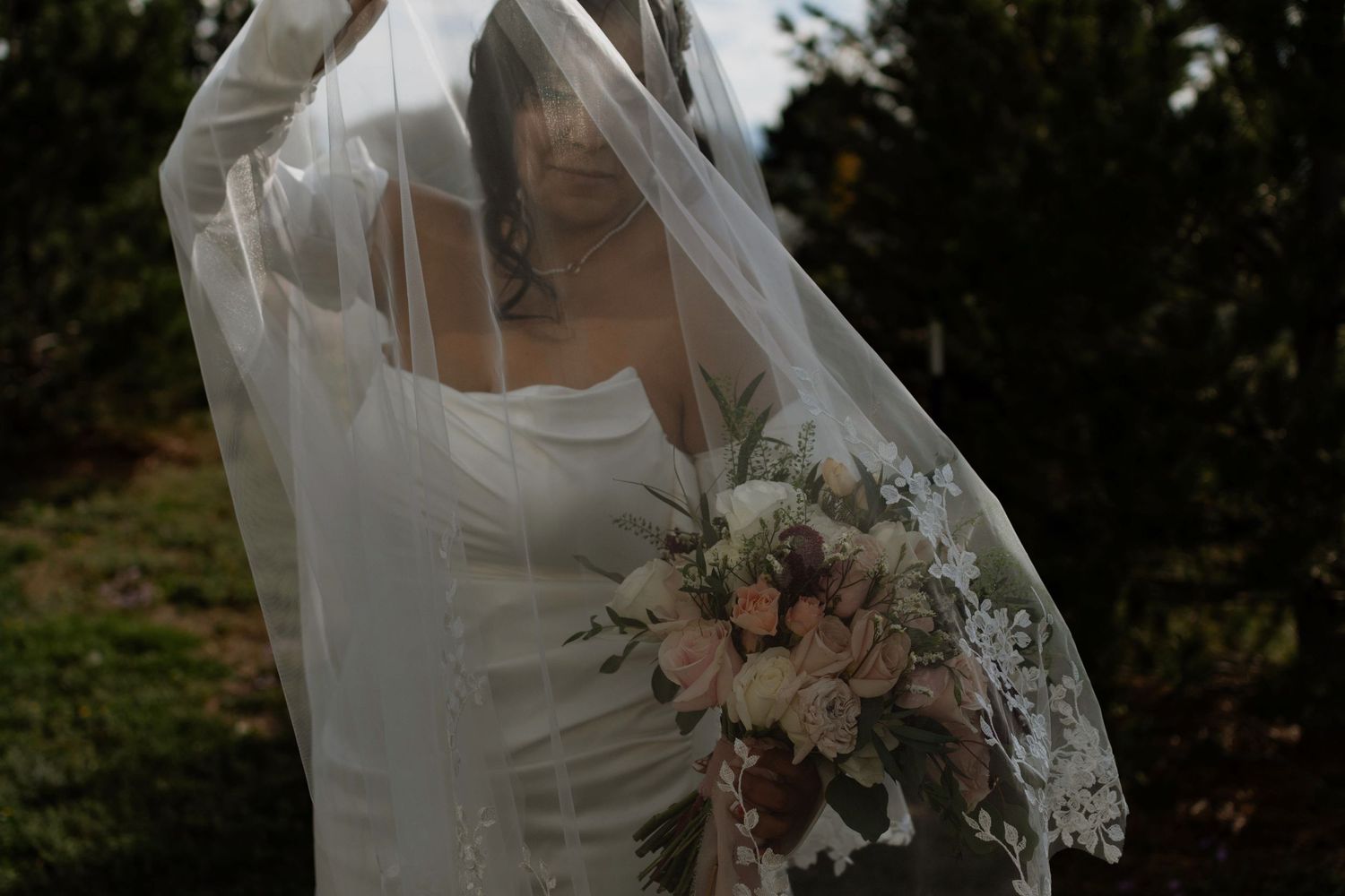 Bride in white gown holding floral bouquet with veil draped overhead outdoors.