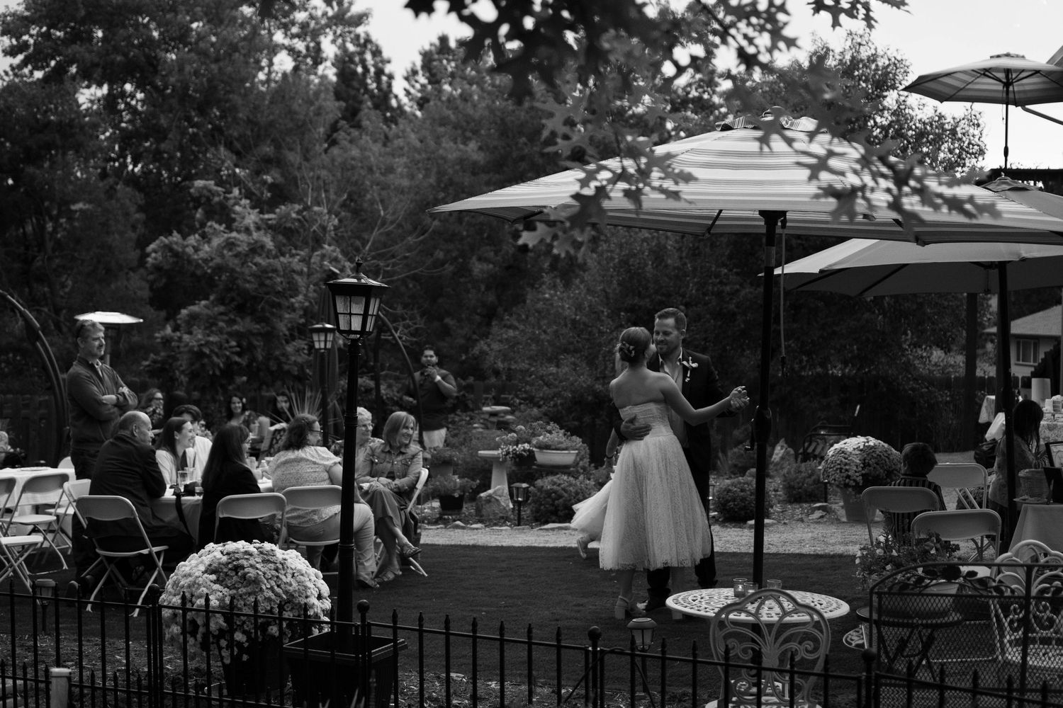 Black and white outdoor wedding reception with couple dancing on patio surrounded by guests, umbrellas, and lush trees.