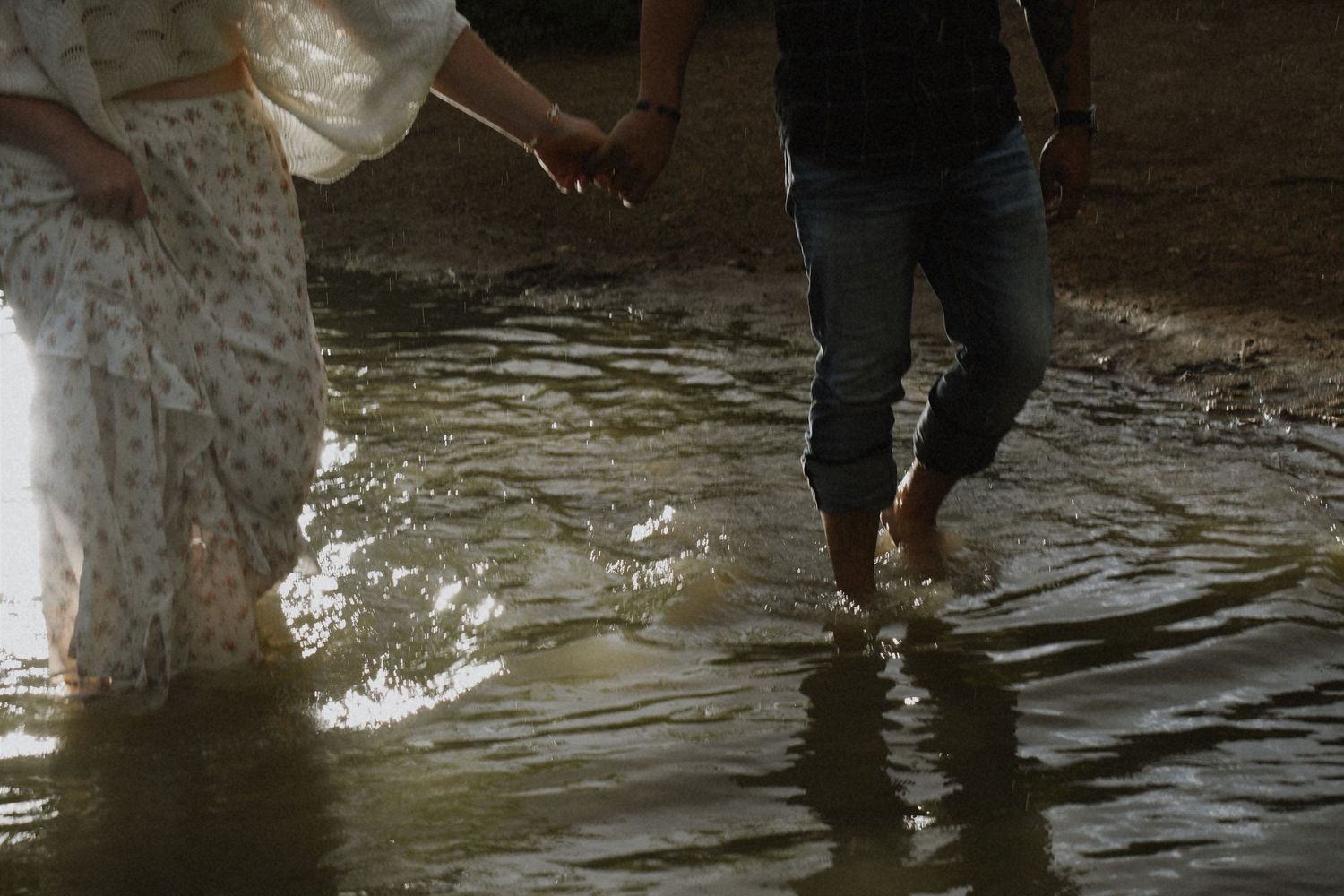 Couple holding hands while wading barefoot through shallow water, wearing casual and white flowy clothing.