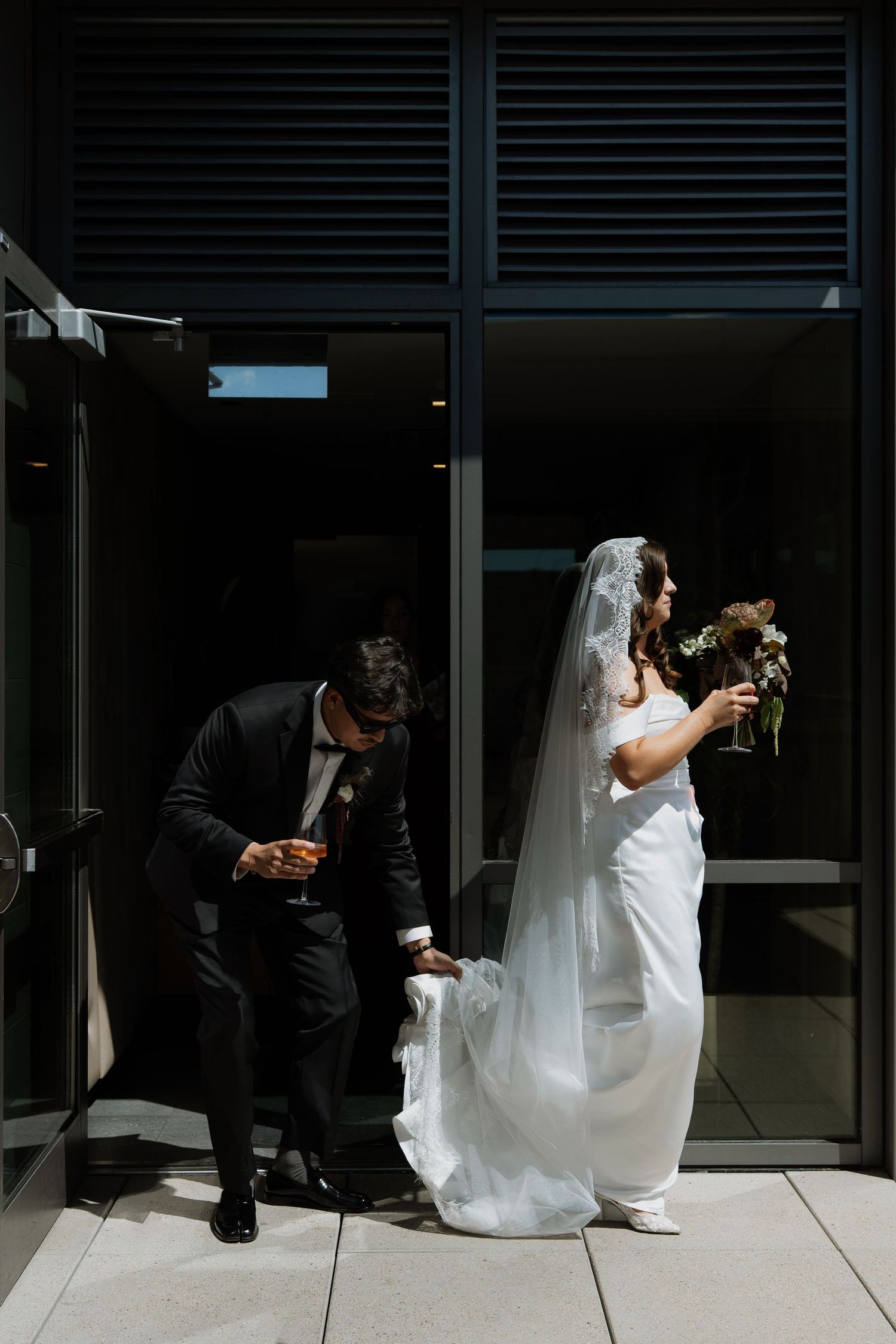 Bride in white gown holding bouquet stands outside modern venue while groom adjusts her train, both holding champagne.