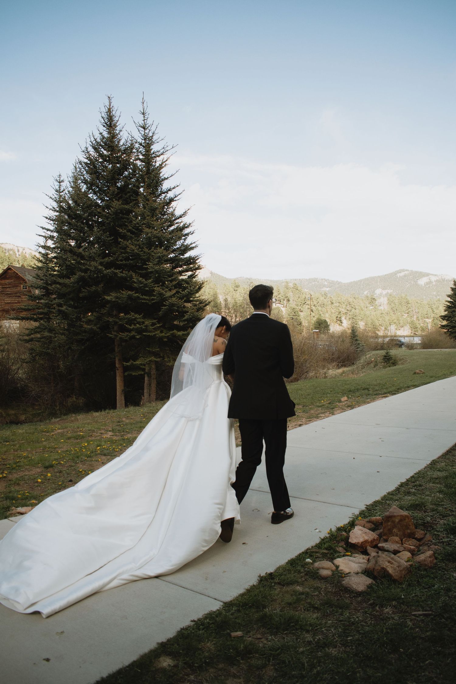 Bride in white gown with long veil walks with groom in black suit along a path with mountain views.