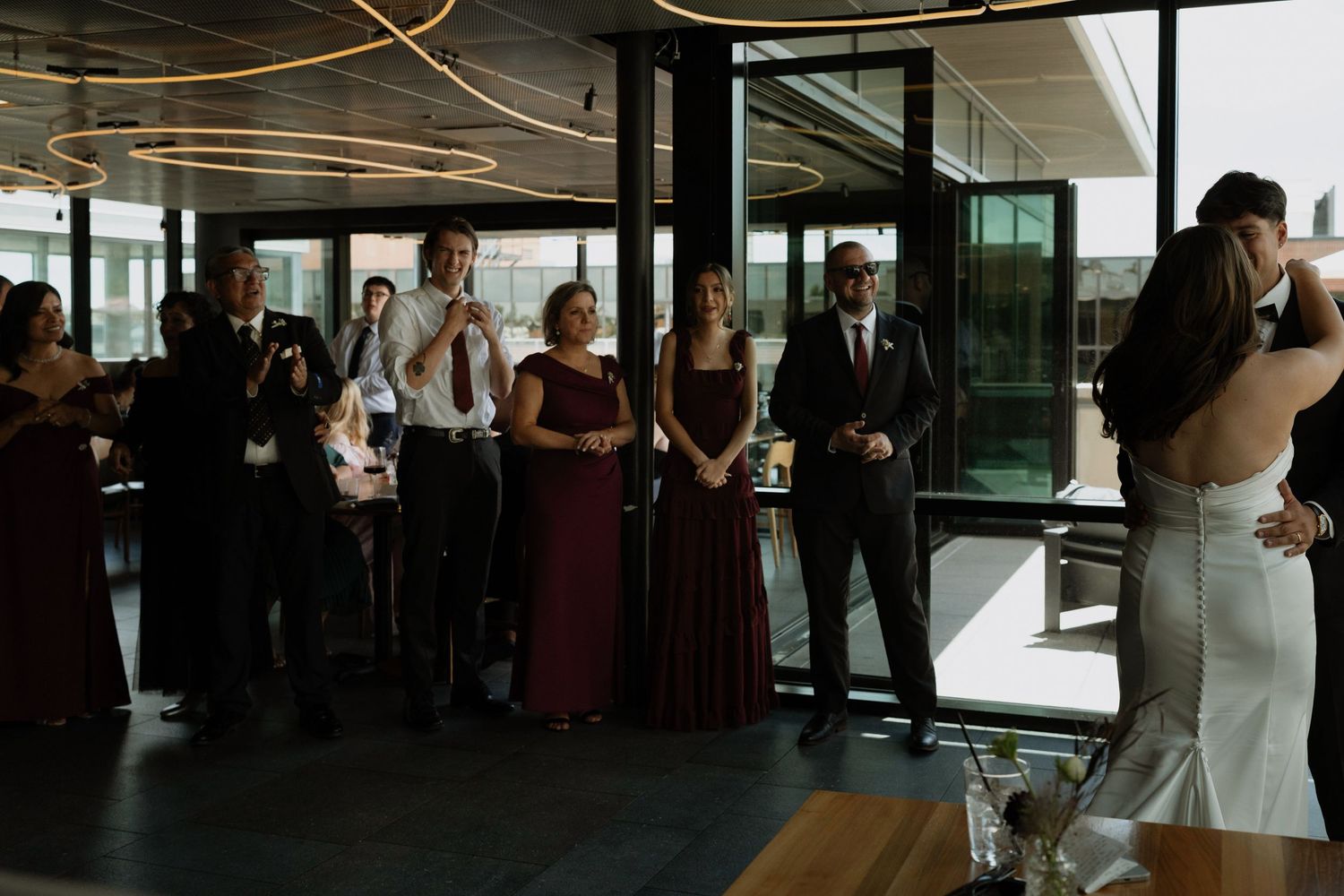 Wedding guests in burgundy dresses watch couple share first dance at modern venue reception.