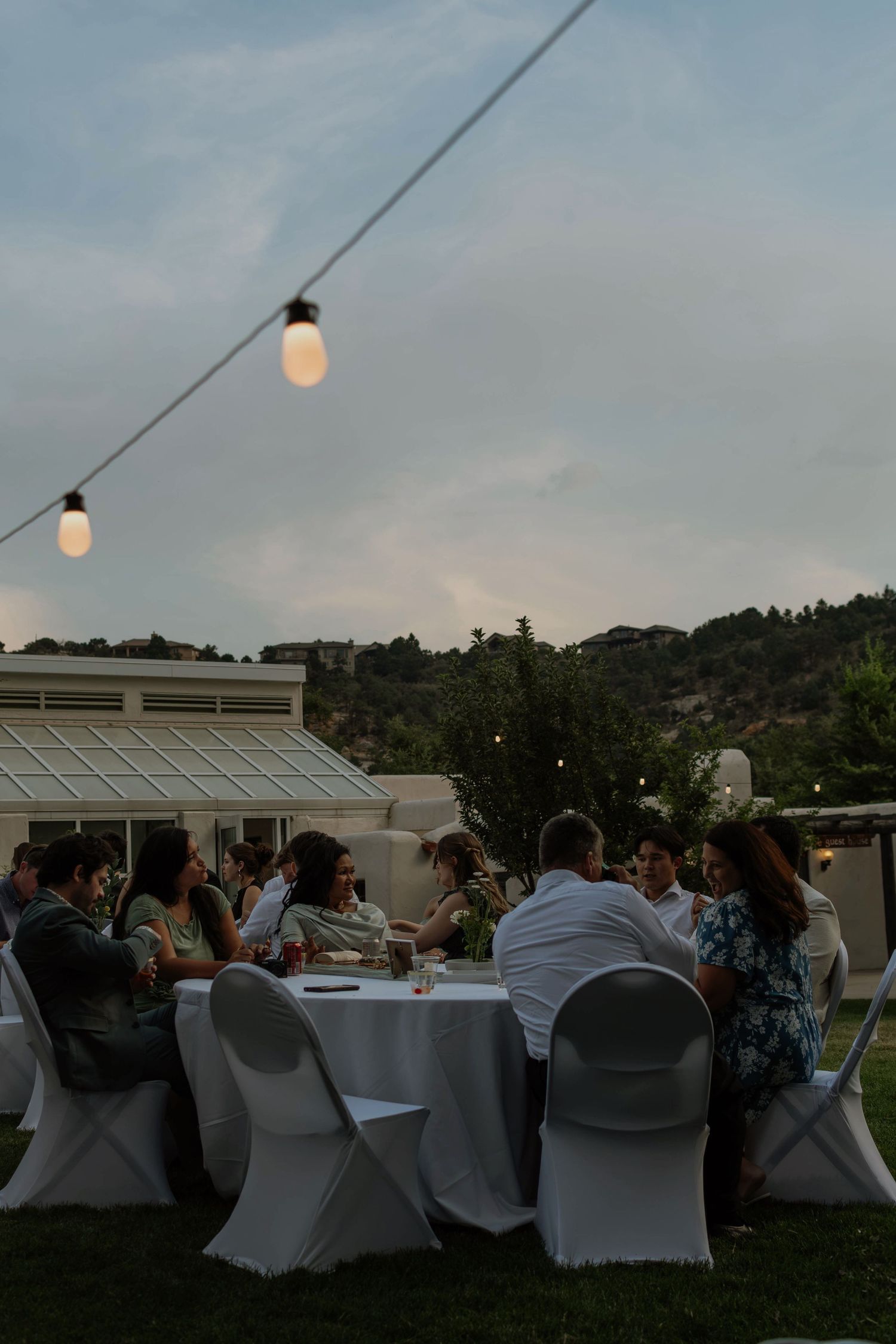 Guests dining outdoors at dusk under string lights at an elegant garden reception with white chairs.