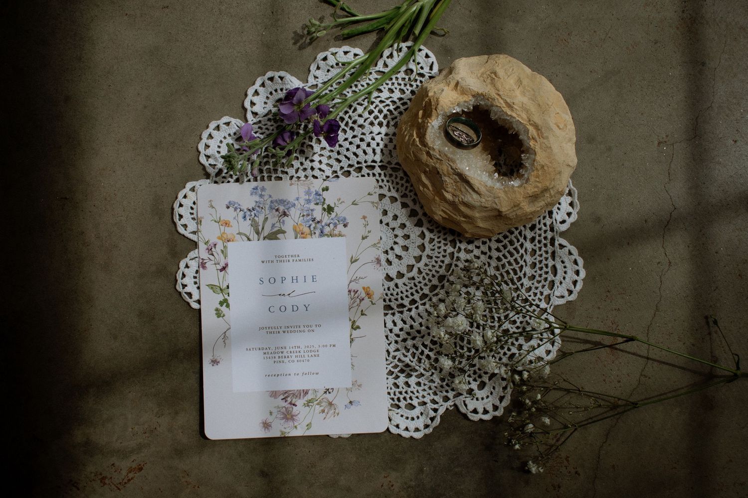 Elegant wedding invitation on lace doily with wildflowers and rustic bread on textured surface.
