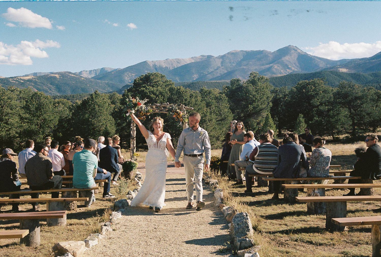 Bride and groom walk down outdoor aisle with Rocky Mountain backdrop at scenic Colorado wedding ceremony.