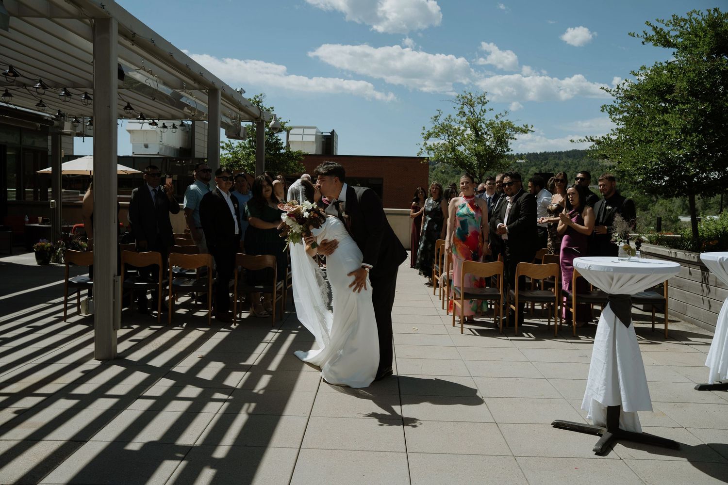 Bride and groom share a romantic kiss on an outdoor patio during their wedding ceremony with guests seated nearby.