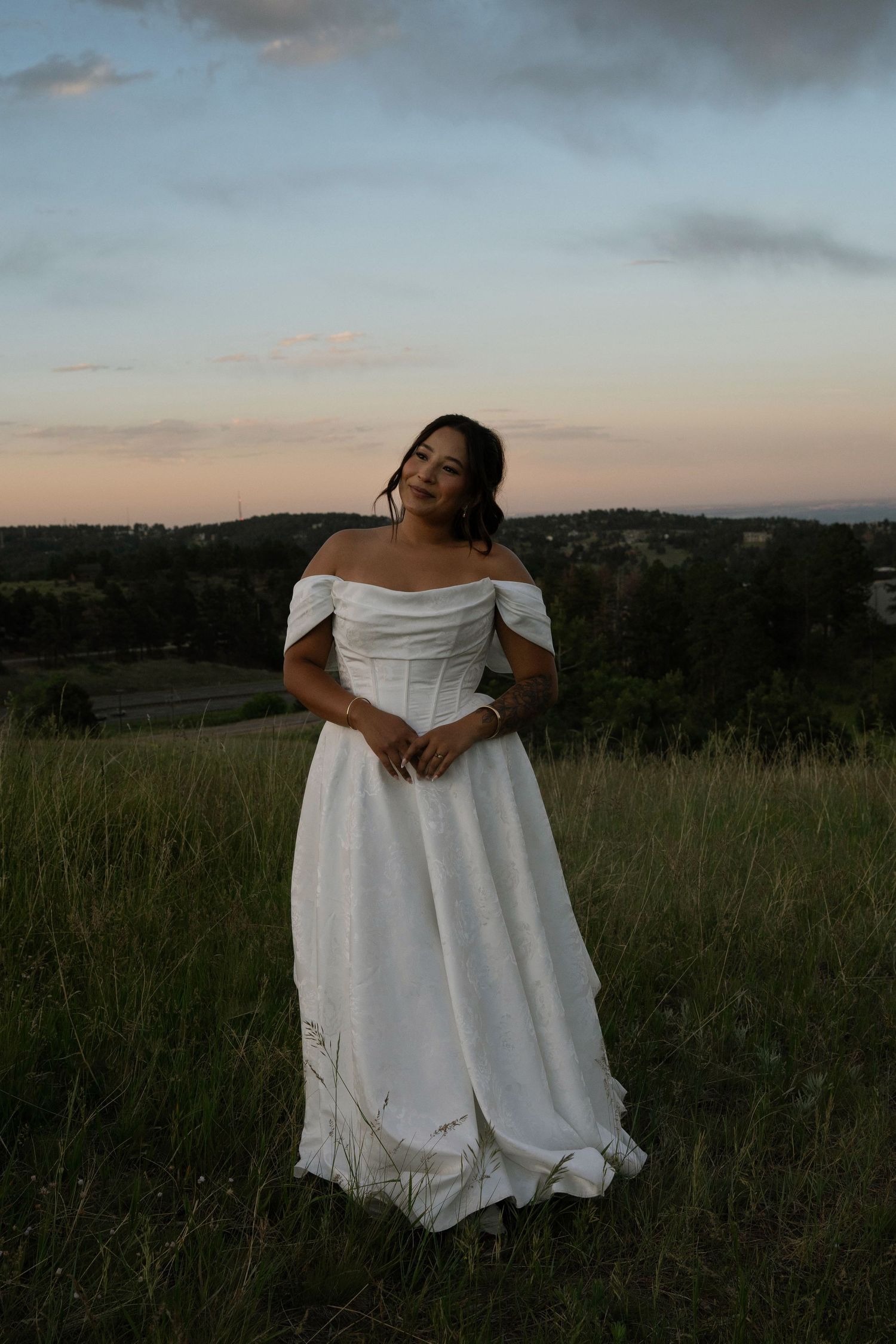 Woman in white off-shoulder wedding gown standing in green meadow at dusk with scenic countryside backdrop.