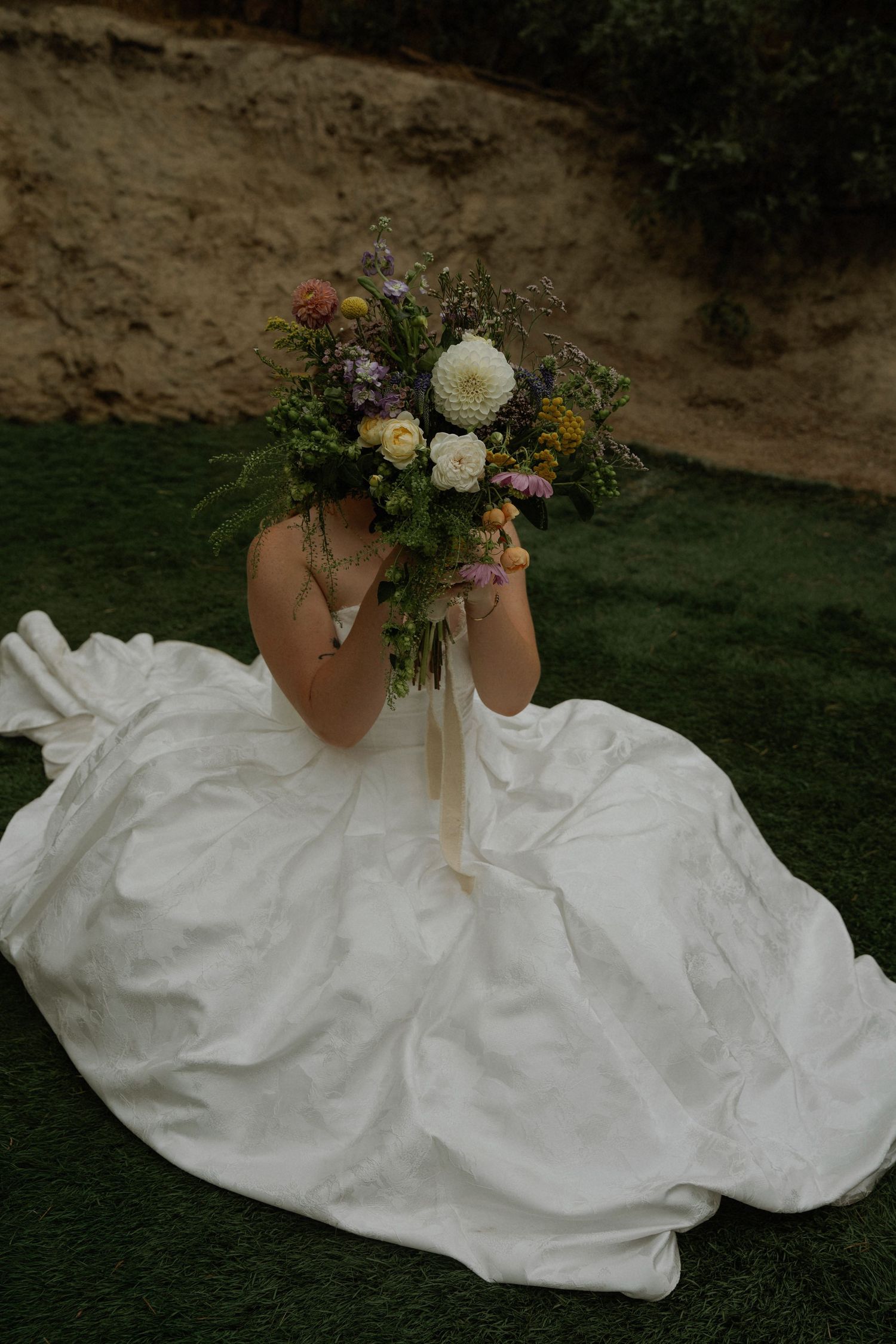Bride in white gown sits on grass holding colorful wildflower bouquet covering her face with ribbon streamers.