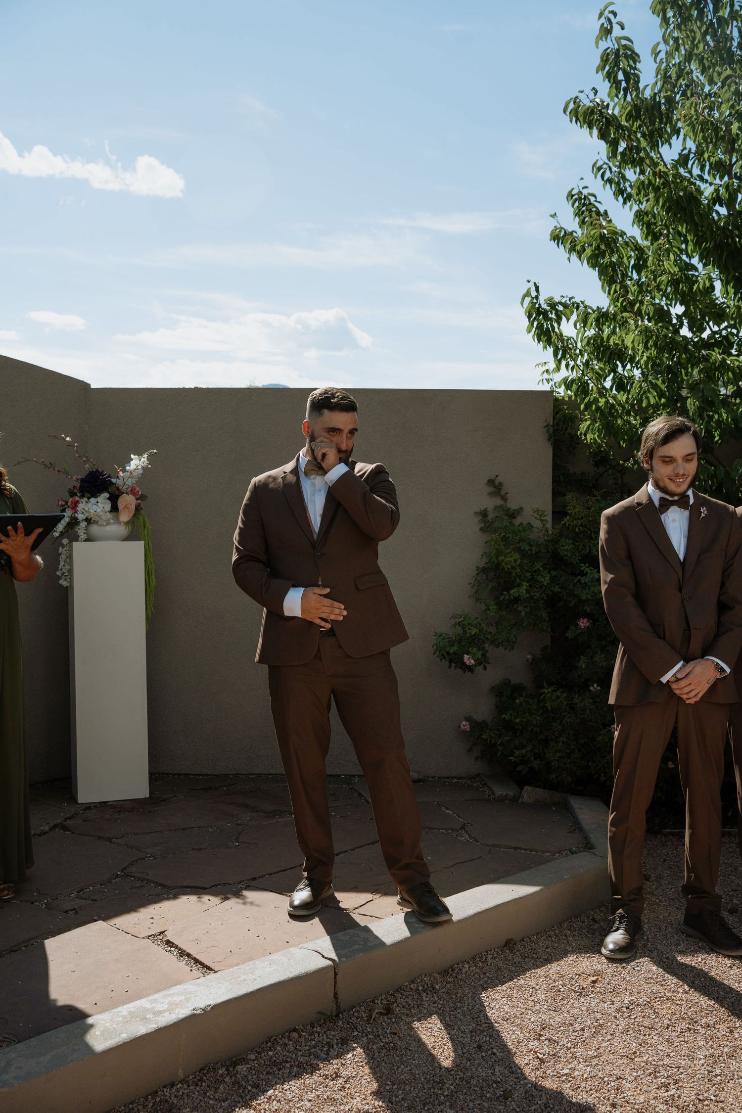 Two groomsmen in brown suits stand outdoors at a wedding ceremony, one emotionally wiping a tear.