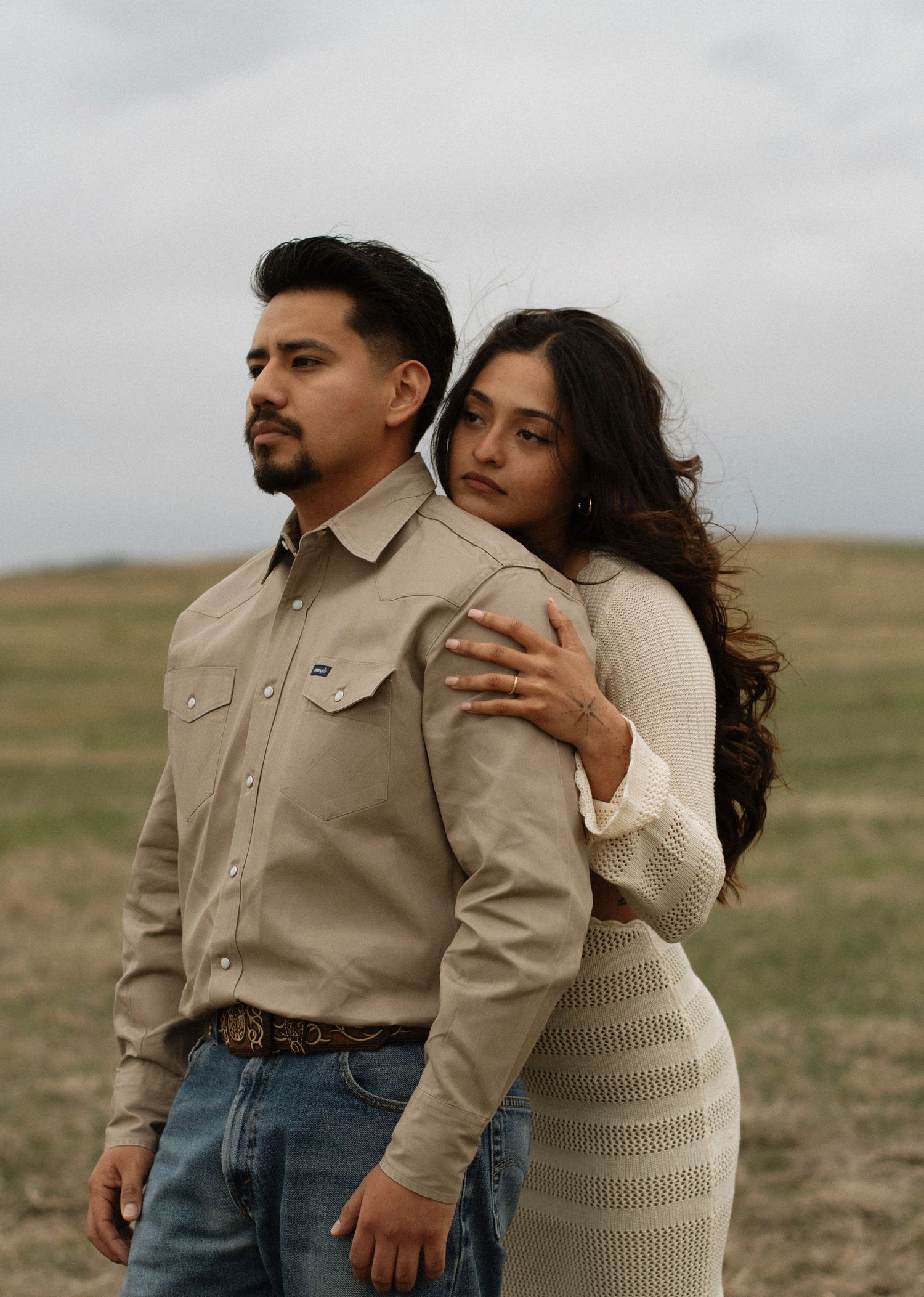 Couple posing in a field, man in beige Wrangler shirt and jeans, woman in cream knit dress embracing him from behind.