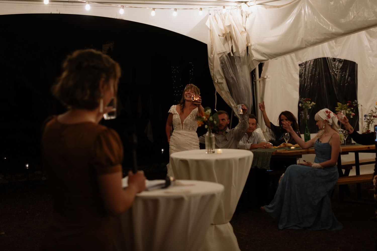 Bride giving a speech at an outdoor wedding reception under a white tent at night with guests seated nearby.