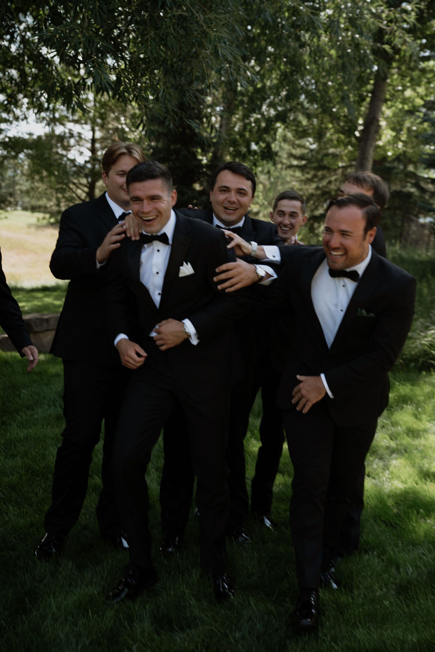 Groom and groomsmen laugh together in black tuxedos outdoors during wedding celebration.