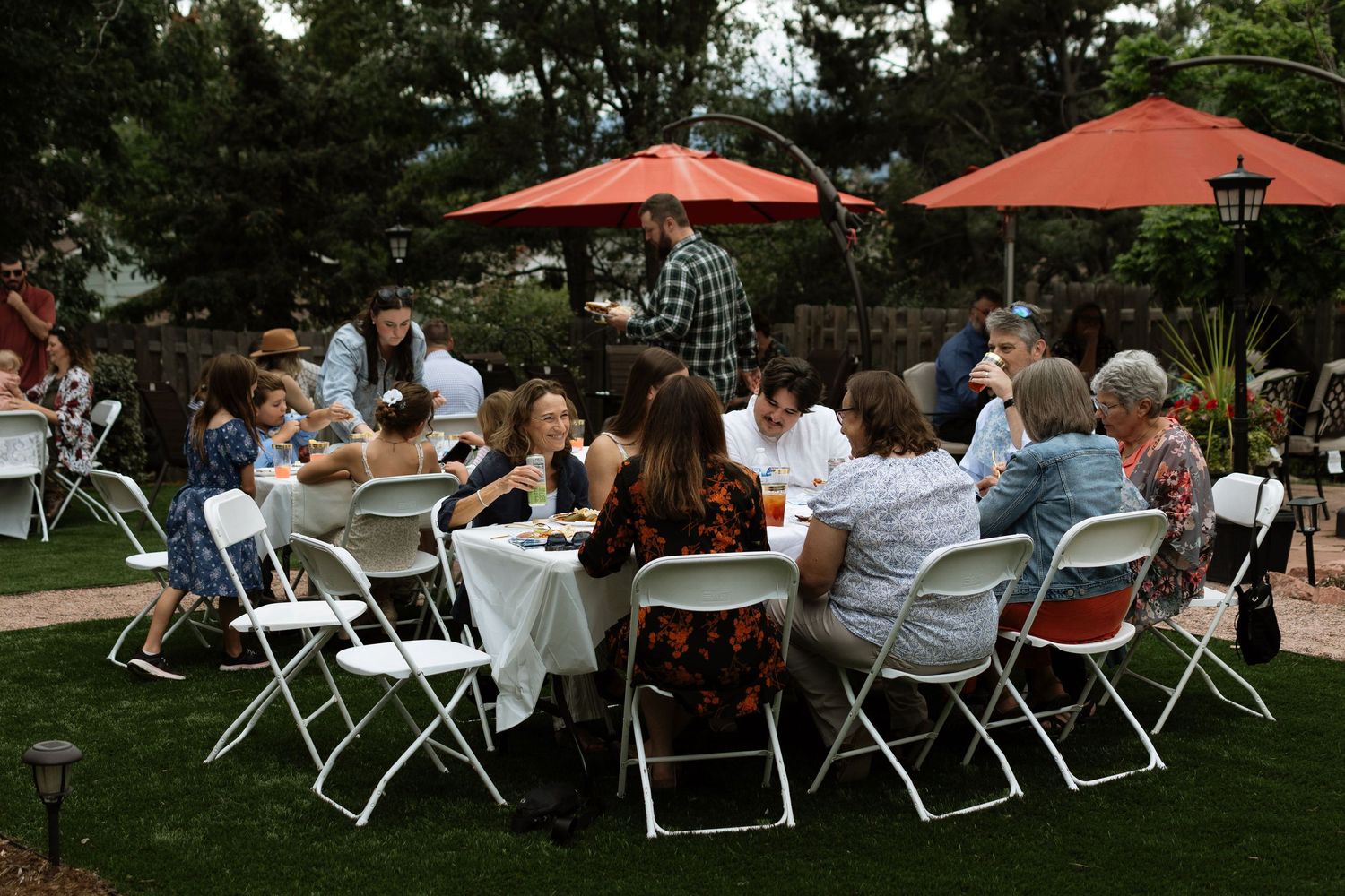 Group of people dining outdoors at round tables with orange umbrellas on a green lawn at a garden party.