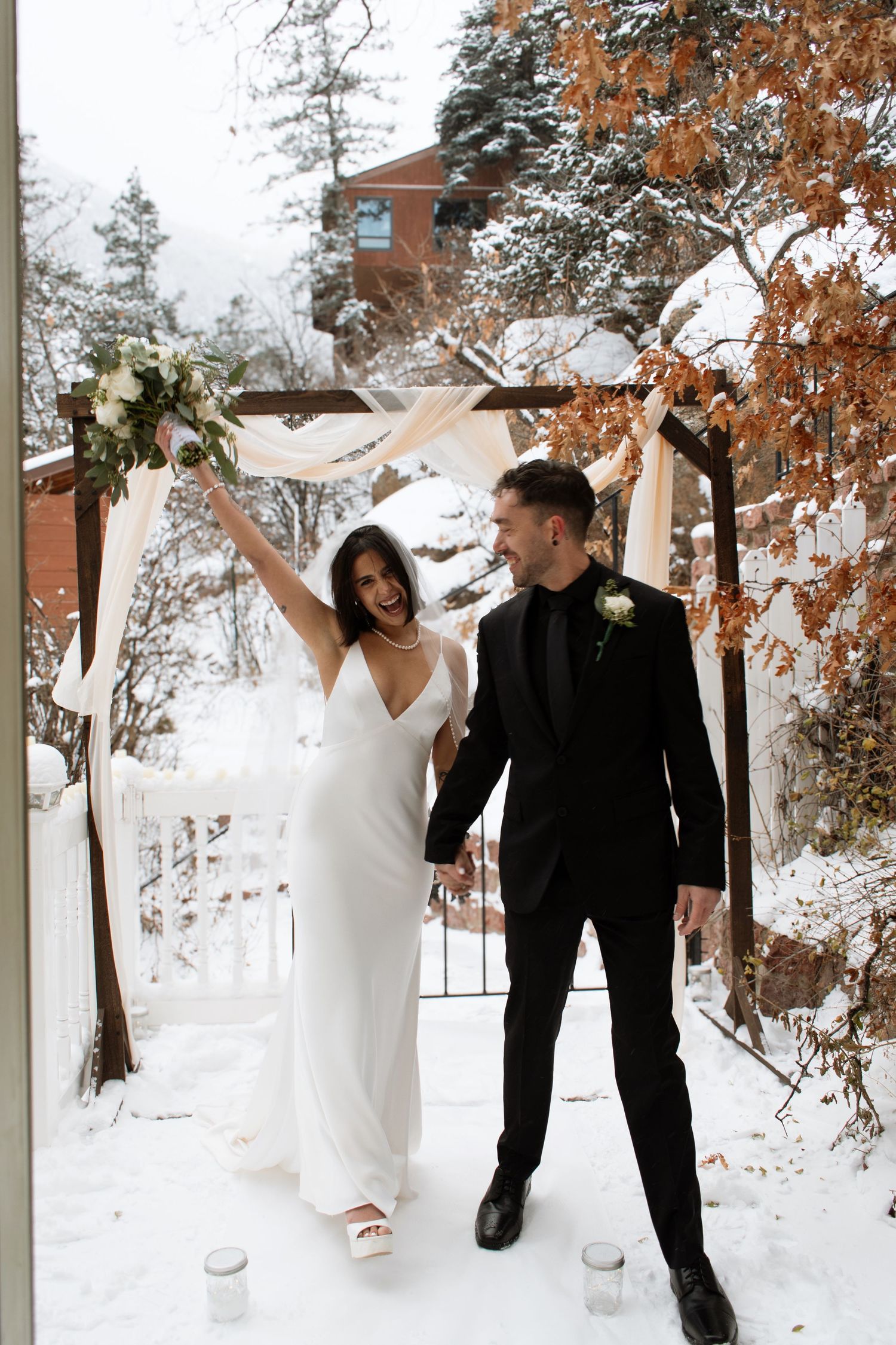Joyful bride in white gown raises bouquet beside groom in black suit under snowy outdoor wedding arch.