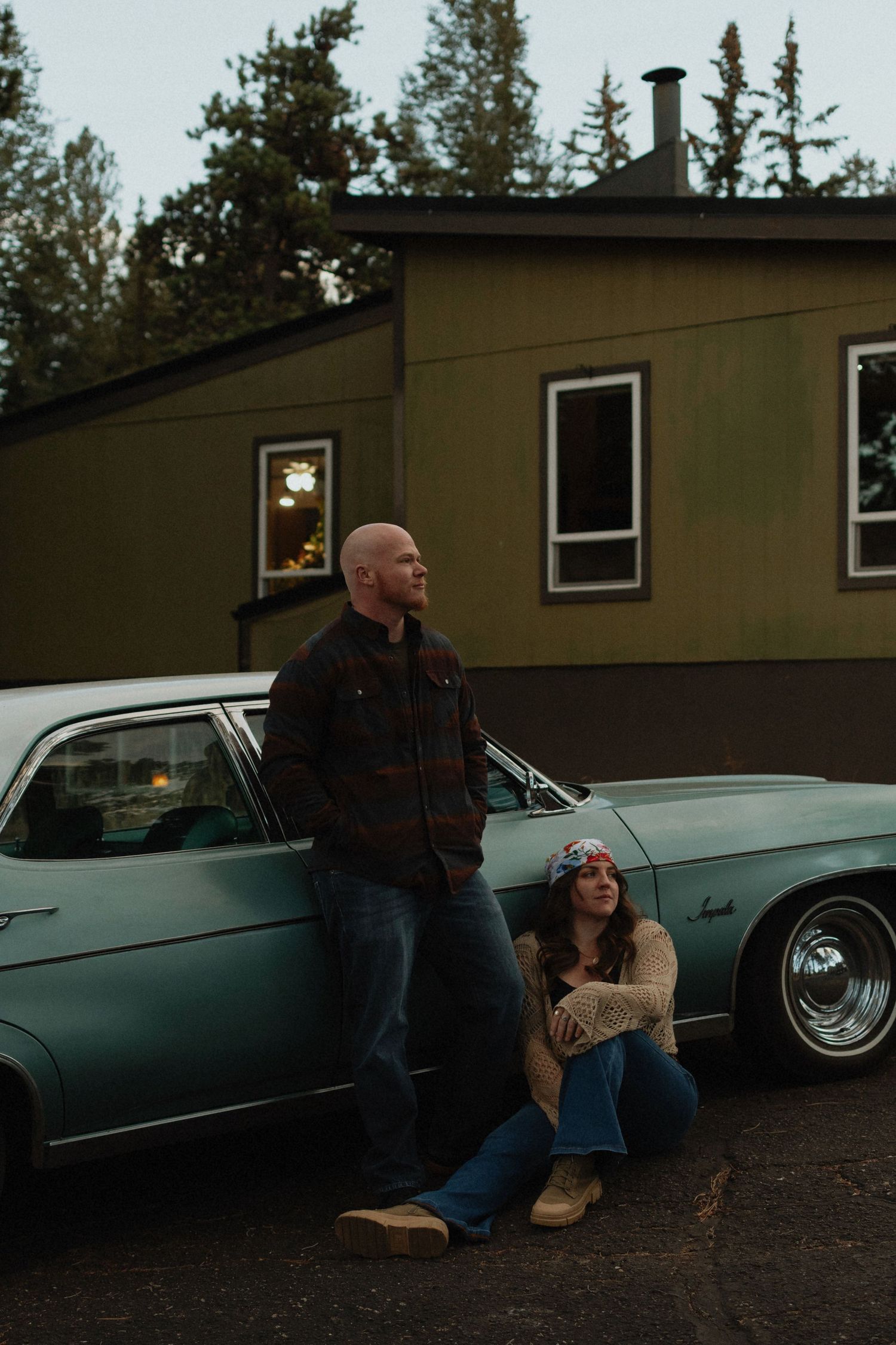 A couple poses beside a vintage teal car at dusk, man standing and woman sitting, with a green house and trees behind them.