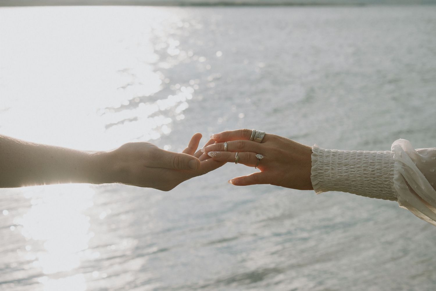 Close-up of two hands gently touching with an engagement ring visible near a shimmering waterfront backdrop.