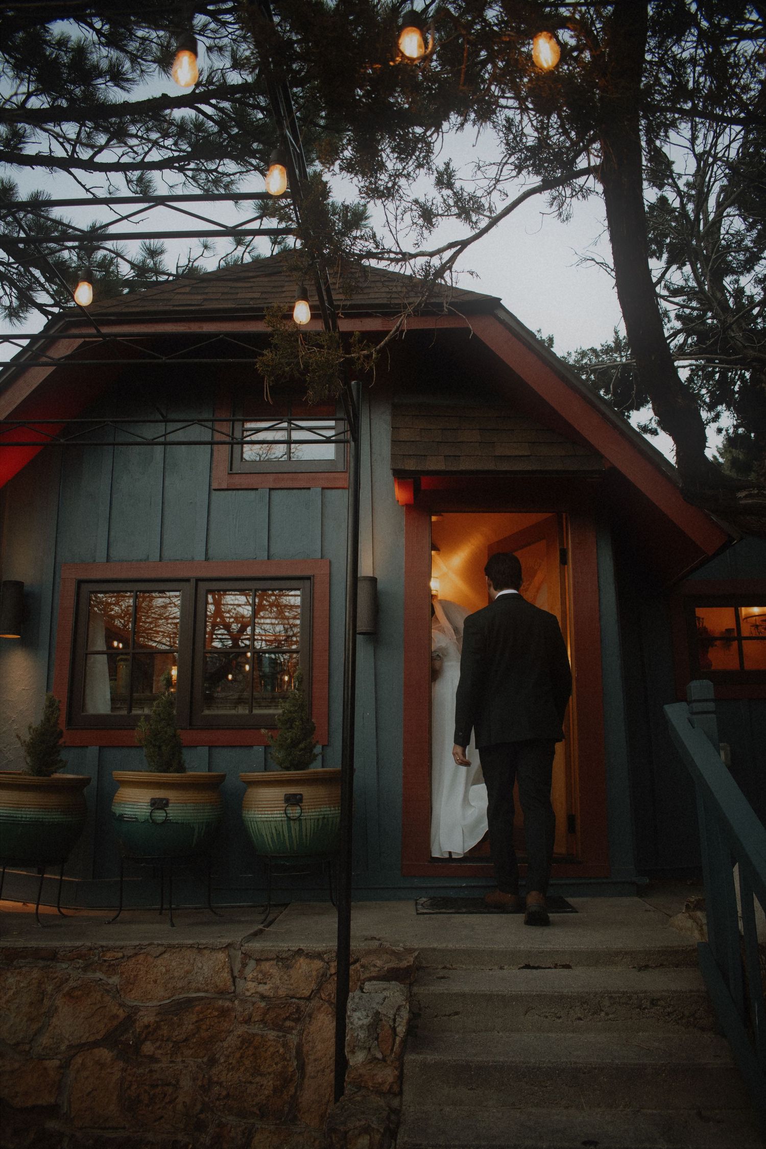 Bride and groom entering a charming blue-gray cabin wedding venue at dusk with warm glowing lights.