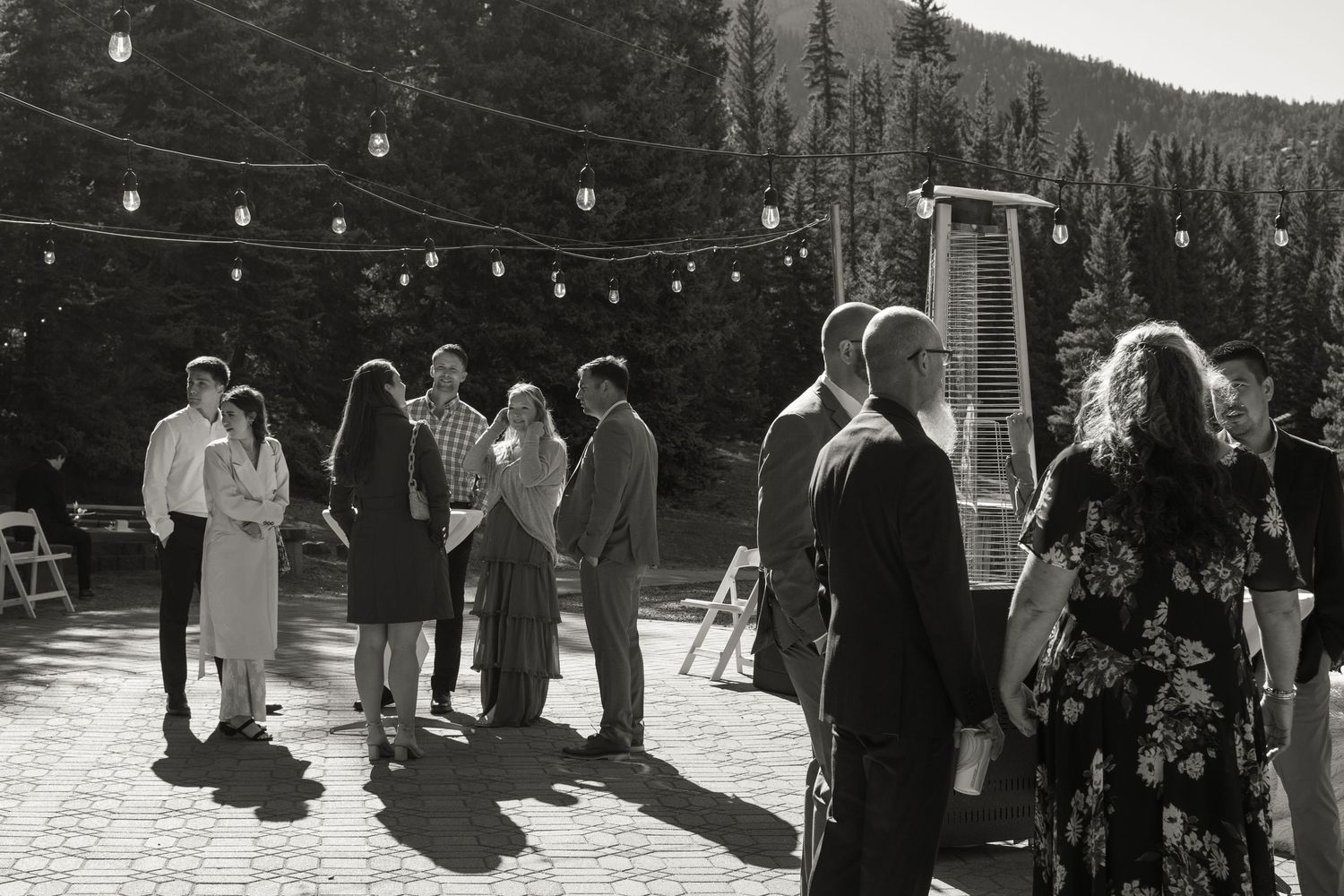 Black and white photo of wedding guests mingling outdoors on a patio with string lights and mountain views.