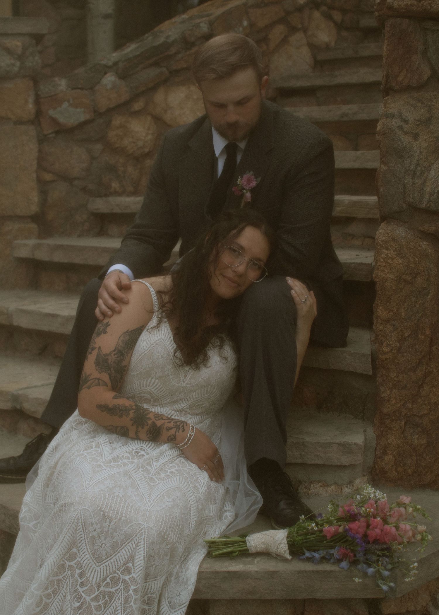 Bride in lace gown with tattoos sits on stone steps leaning against groom in dark suit with floral boutonniere.