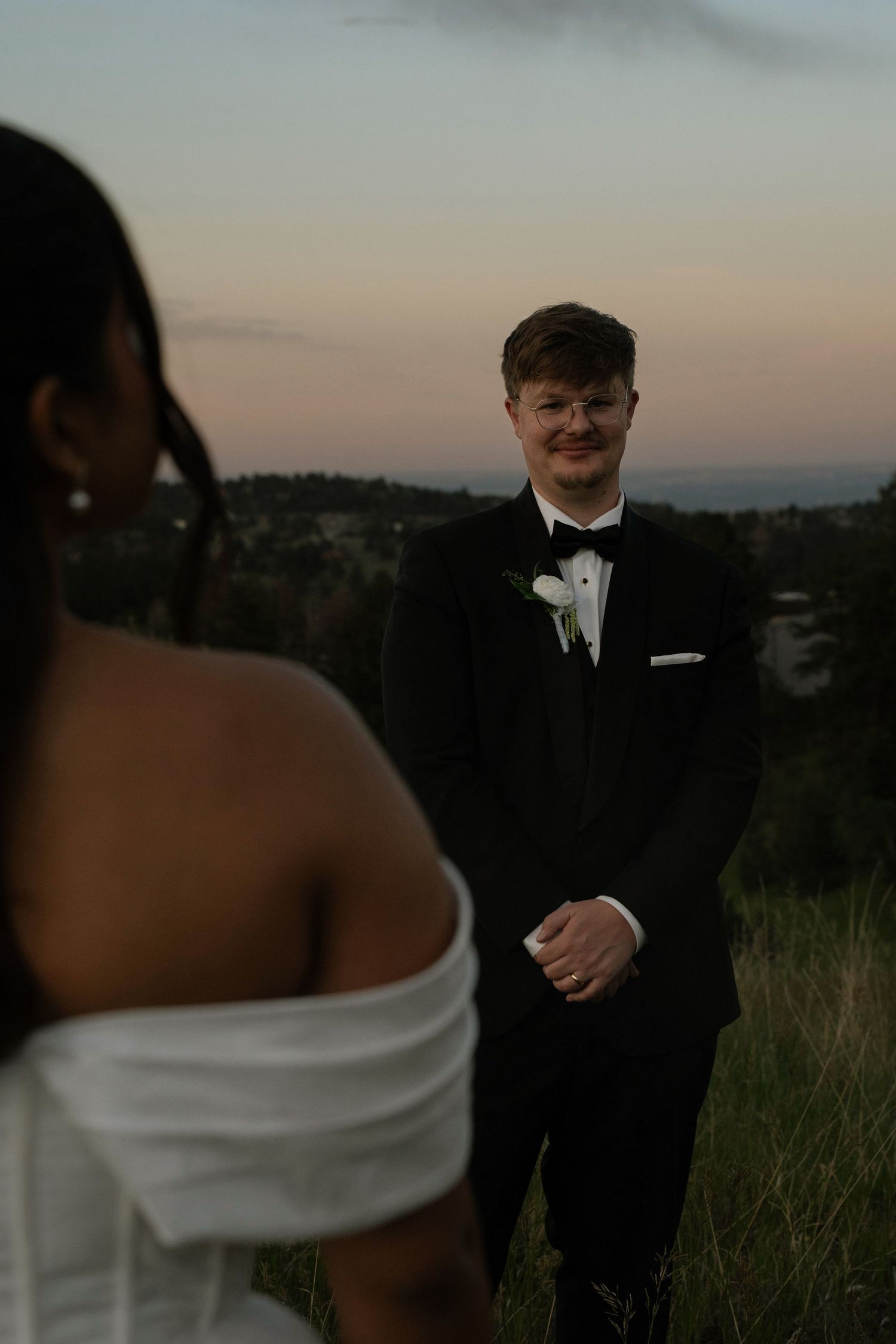 Groom in black tuxedo smiling at bride in white off-shoulder gown during outdoor sunset wedding ceremony.